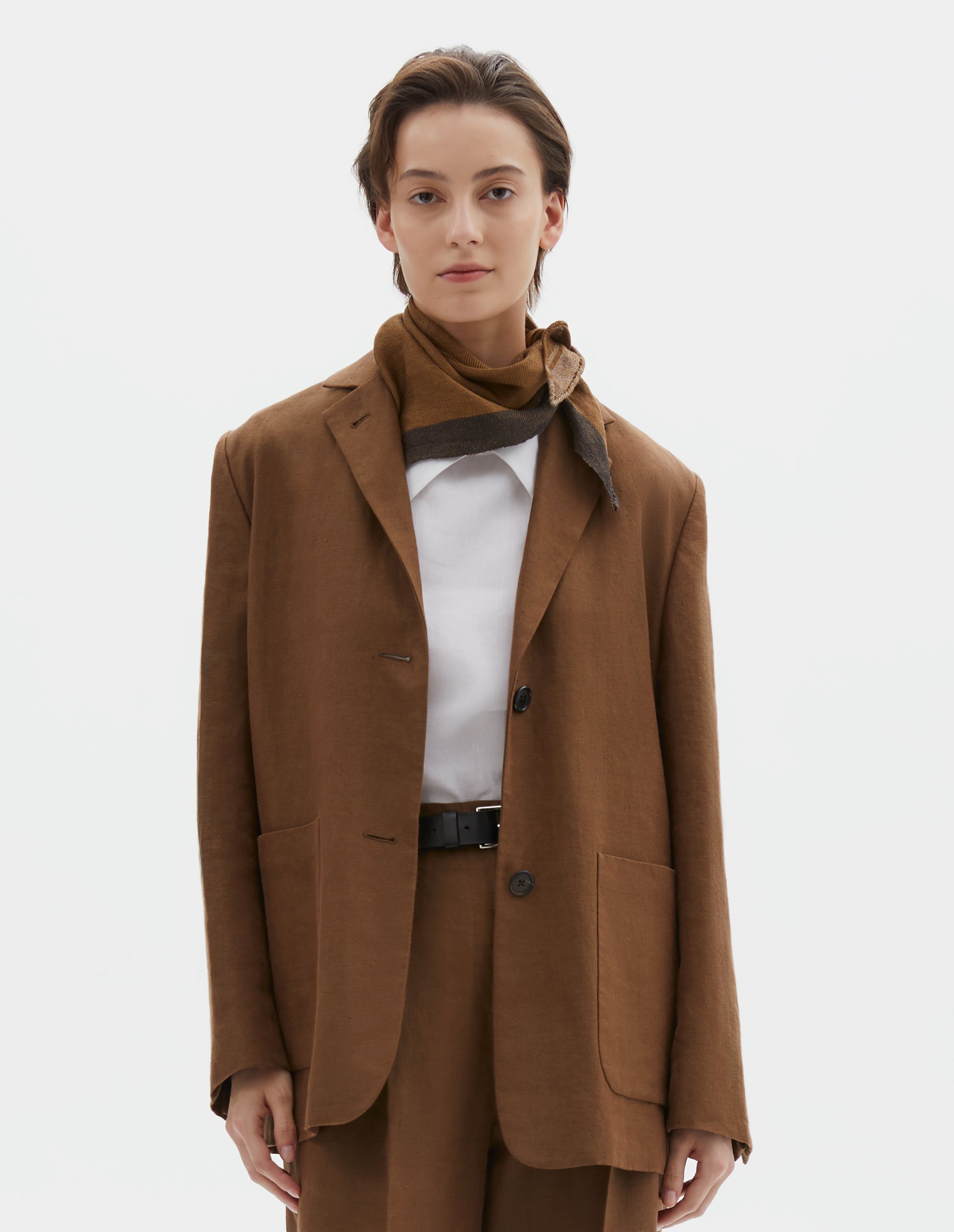 Cropped body image, front view of model wearing a brown, white and black knotted scout scarf. Styled with a brown linen blazer, with dropped shoulders, large patch pockets and horn button closure and cuffs, over white cotton vest, with brown linen trousers.