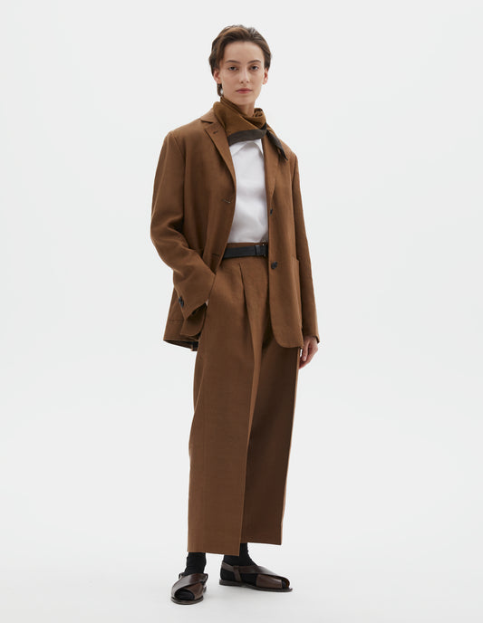 Full body image, front view of model wearing brown blazer with dropped shoulders, large patch pockets and horn button closure and cuffs.  Styled with brown scout scarf, white top, black belt, matching brown trousers and black socks with dark brown leather sandals. 