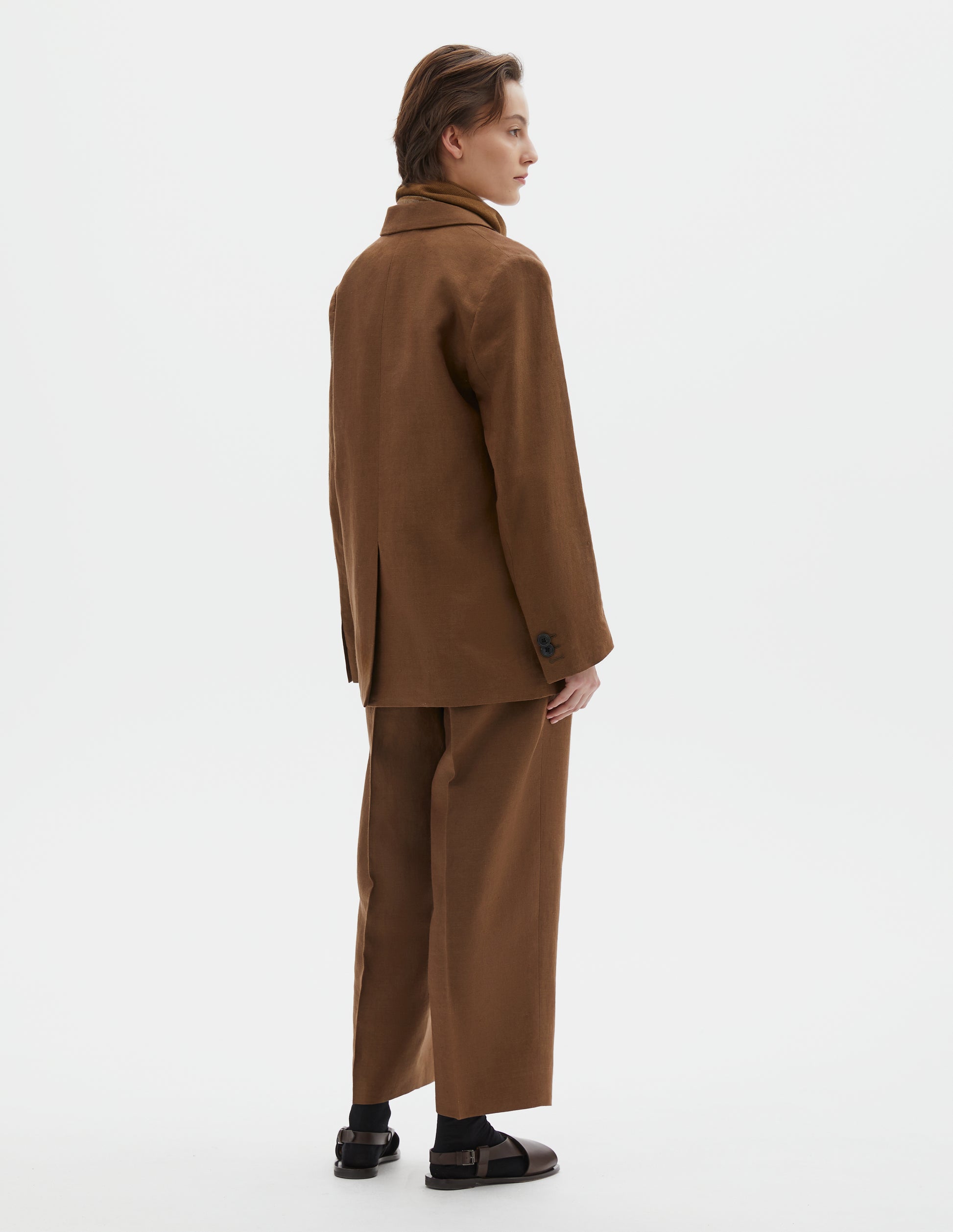 Full body image, back view of model wearing brown blazer with horn button cuffs and back vent. Styled with brown scout scarf, white top, black belt, matching brown trousers and black socks with dark brown leather sandals.