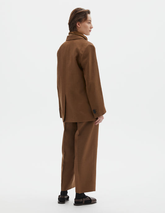 Full body image, back view of model wearing brown blazer with horn button cuffs and back vent. Styled with brown scout scarf, white top, black belt, matching brown trousers and black socks with dark brown leather sandals.