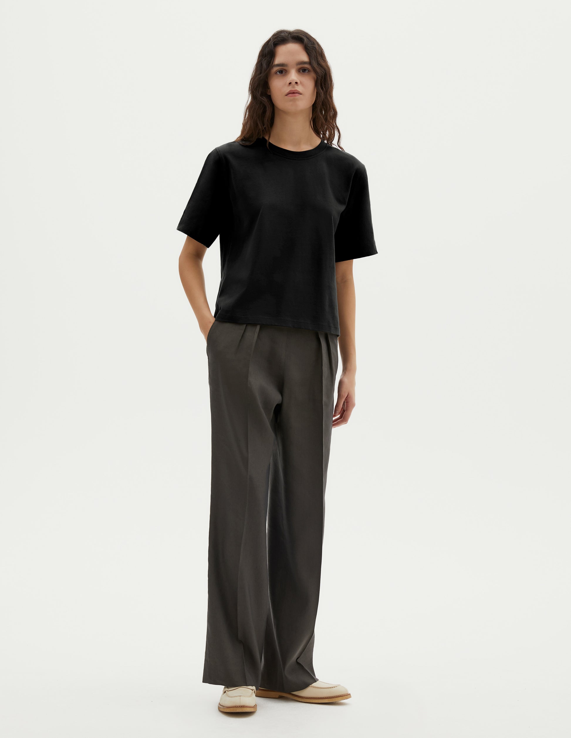 Full body image, front view of model wearing black cotton short sleeve t-shirt with boxy cut. Styled with green pleated trousers and beige suede loafer. 