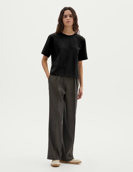 Full body image, front view of model wearing black cotton short sleeve t-shirt with boxy cut. Styled with green pleated trousers and beige suede loafer. 