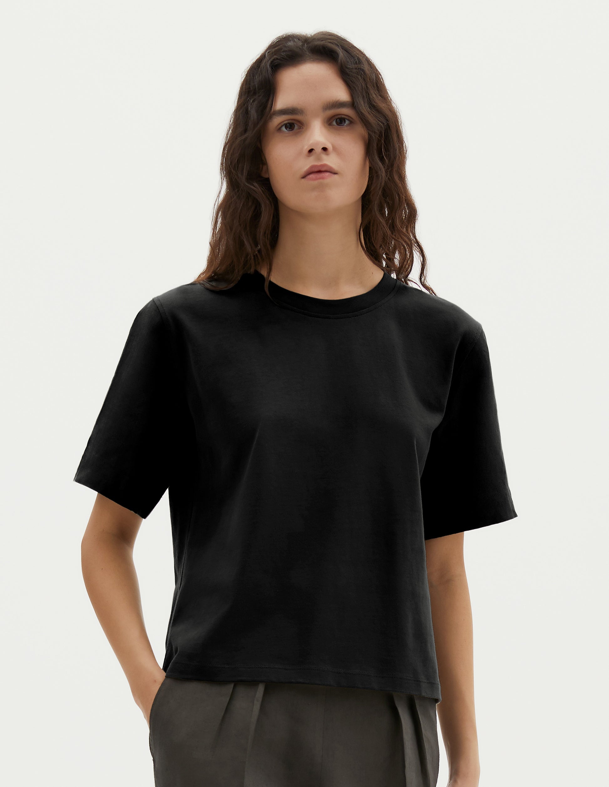 Cropped body image, front view of model wearing black cotton short sleeve t-shirt with boxy cut. Styled with green pleated trousers.