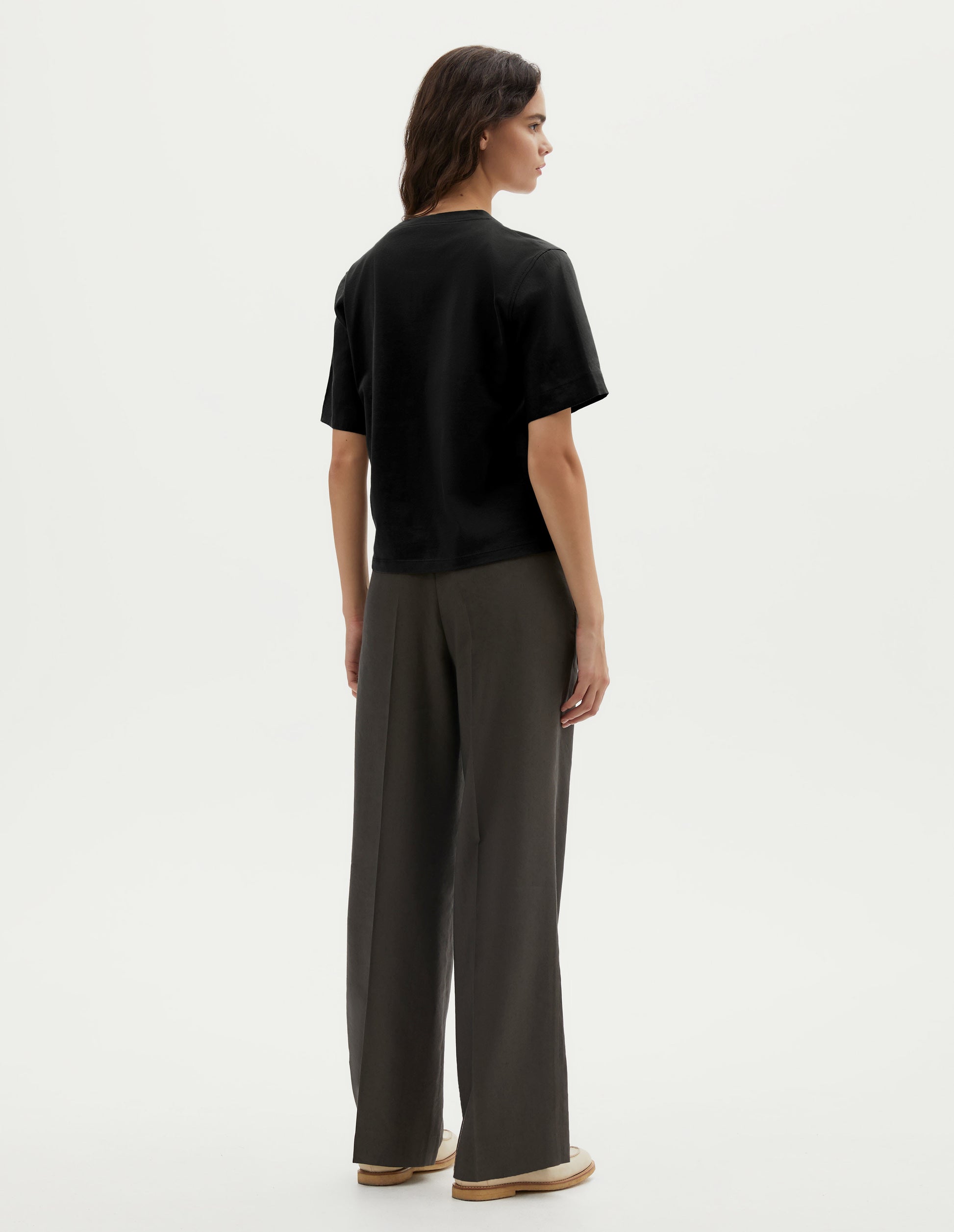 Full body image, back view of model wearing black cotton short sleeve t-shirt with boxy cut. Styled with green pleated trousers and beige suede loafers. 