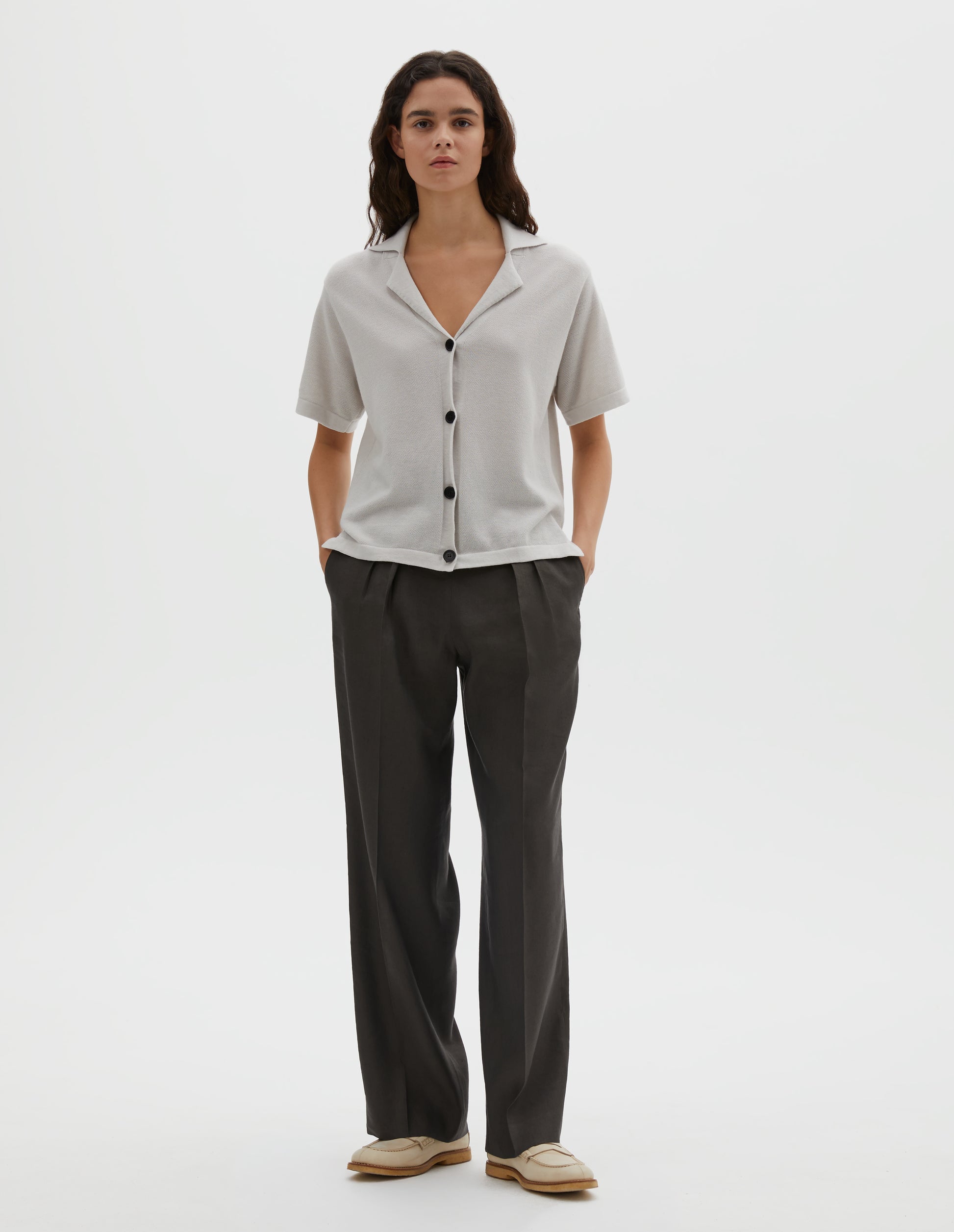 Full body image, front view of model wearing off white knitted cotton short sleeve shirt, with fine gauge rib collar, placket, cuffs and side split hem with horn buttons. Styled over brownish green pleated trousers with cream shoes.