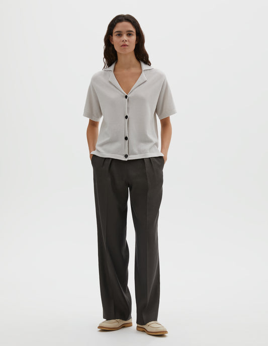 Full body image, front view of model wearing off white knitted cotton short sleeve shirt, with fine gauge rib collar, placket, cuffs and side split hem with horn buttons. Styled over brownish green pleated trousers with cream shoes.