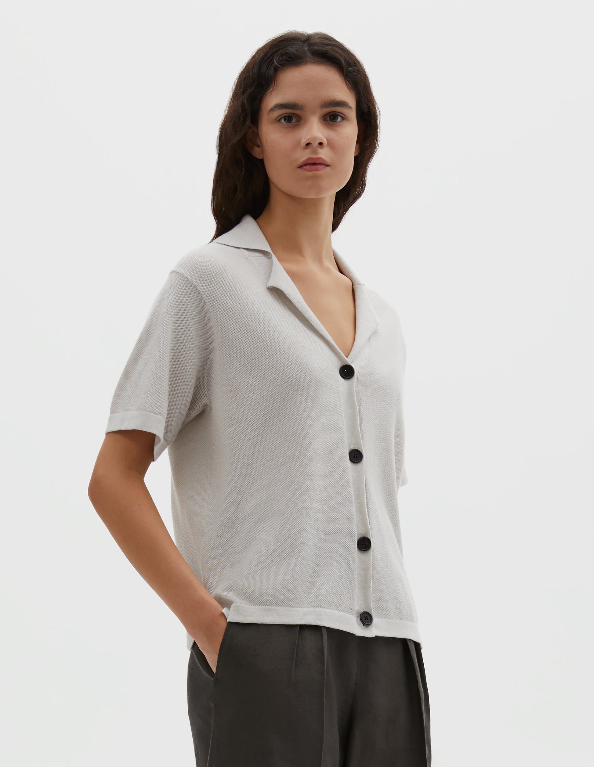 Cropped body image, front view of model wearing off white knitted cotton short sleeve shirt, fine gauge rib collar, placket, cuffs and side split hem with dark horn buttons. Styled over brownish green pleated trousers.