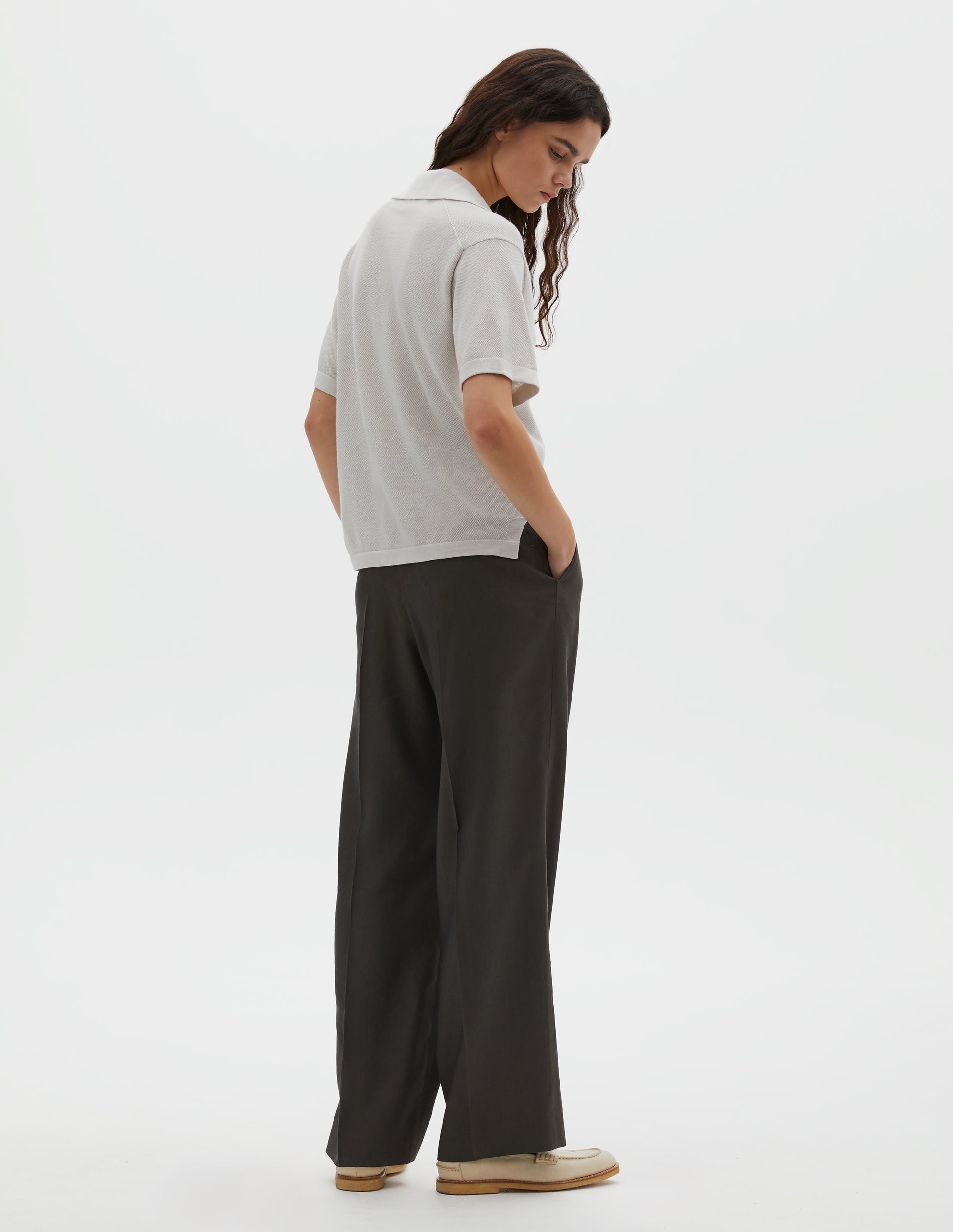 Full body image, back view of model wearing off white knitted cotton short sleeve shirt, with fine gauge rib collar, placket, cuffs and side split hem. Styled over brownish green pleated trousers with cream shoes.