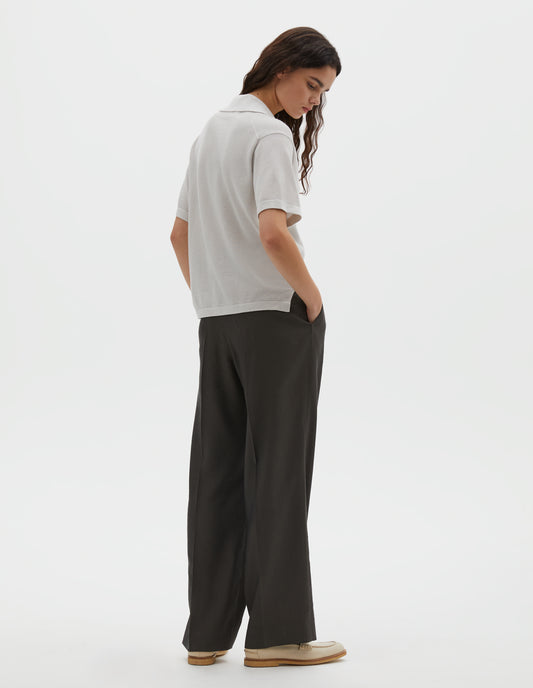 Full body image, back view of model wearing off white knitted cotton short sleeve shirt, with fine gauge rib collar, placket, cuffs and side split hem. Styled over brownish green pleated trousers with cream shoes.