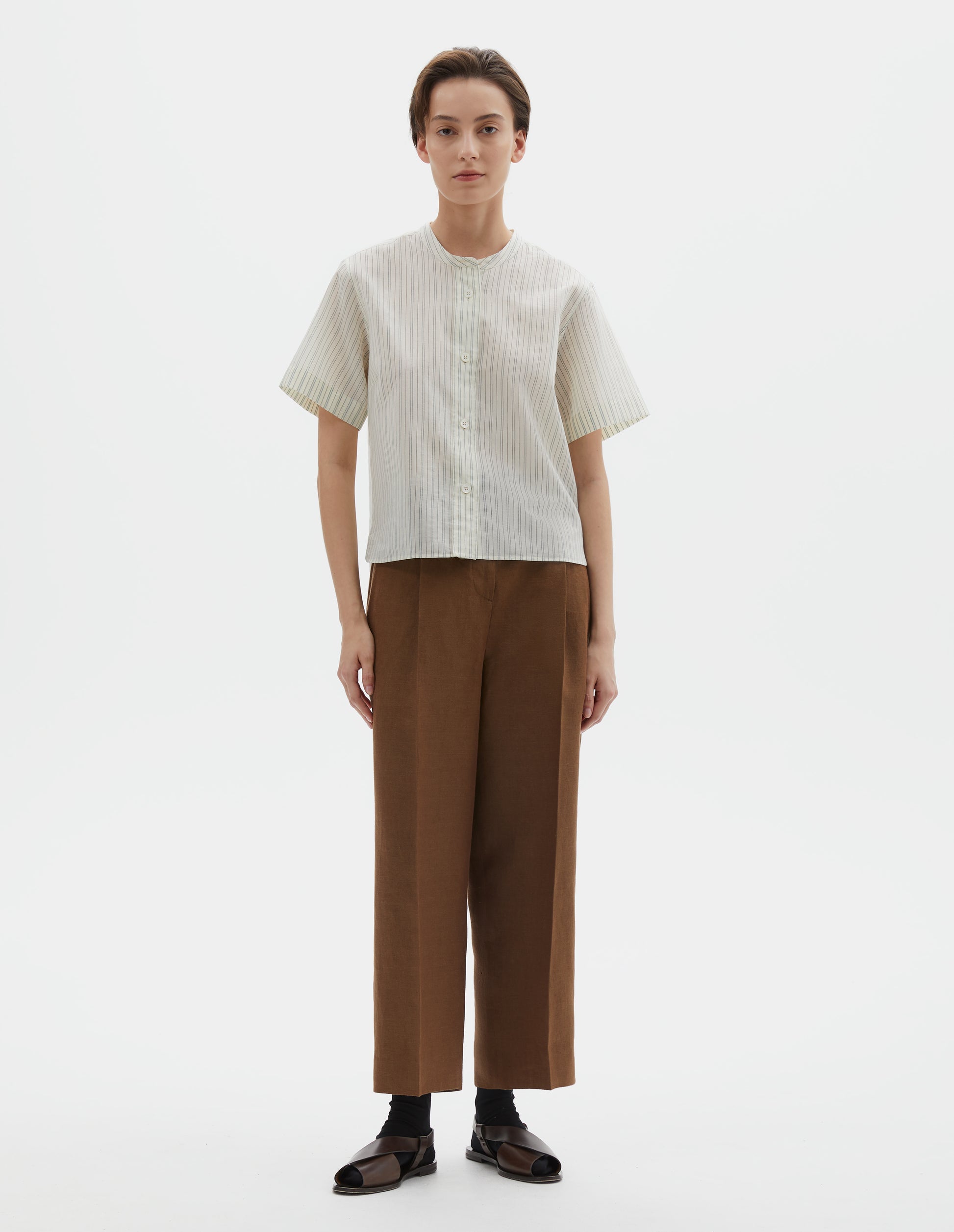 Full body image, front view of model wearing regular cut cropped striped beige and light blue collarless shirt with short sleeves, side split stepped hem and mother of pearl buttons on raised placket. Styled with brown trousers, black socks and dark brown leather sandals.