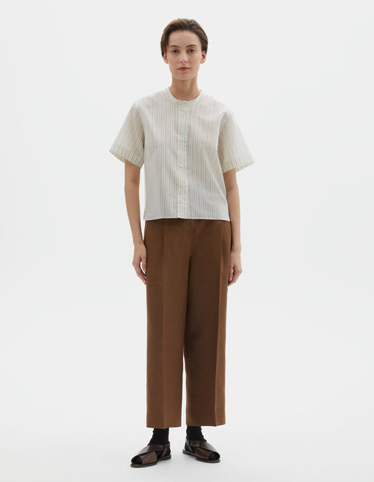 Full body image, front view of model wearing regular cut cropped striped beige and light blue collarless shirt with short sleeves, side split stepped hem and mother of pearl buttons on raised placket. Styled with brown trousers, black socks and dark brown leather sandals.