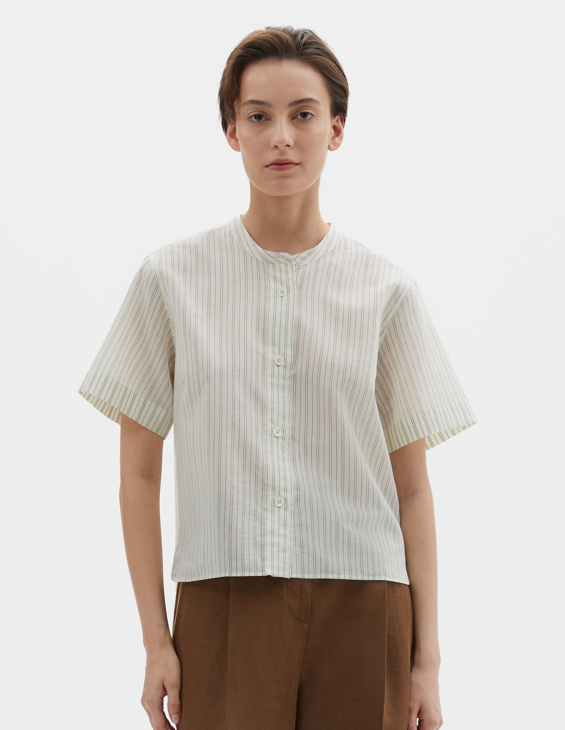 Cropped body image, front view of model wearing regular cut cropped striped beige and light blue collarless shirt with short sleeves, side split stepped hem and mother of pearl buttons on raised placket. Styled with brown trousers.