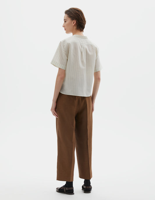 Full body image, back view of model wearing regular cut cropped striped beige and light blue collarless shirt with short sleeves, side split stepped hem, box pleat and hanger loop detail on back yoke, and buttonhole detailing on collar band. Styled with brown trousers, black socks and dark brown leather sandals.