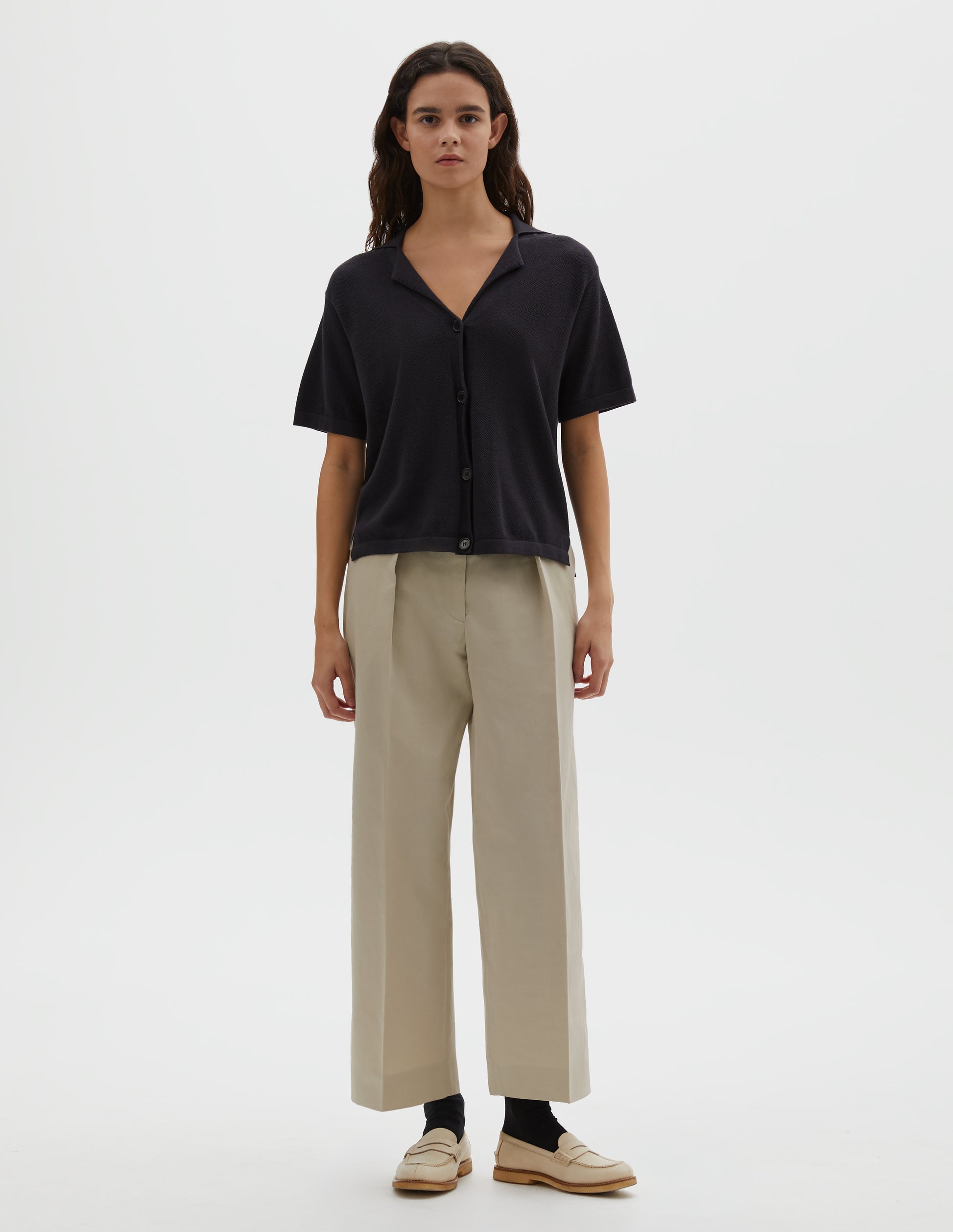 Full body image, front view of model wearing black knitted cotton short sleeve shirt, with fine gauge rib collar, placket, cuffs and side split hem with horn buttons. Styled over cream pleated trousers with black socks and cream shoes.