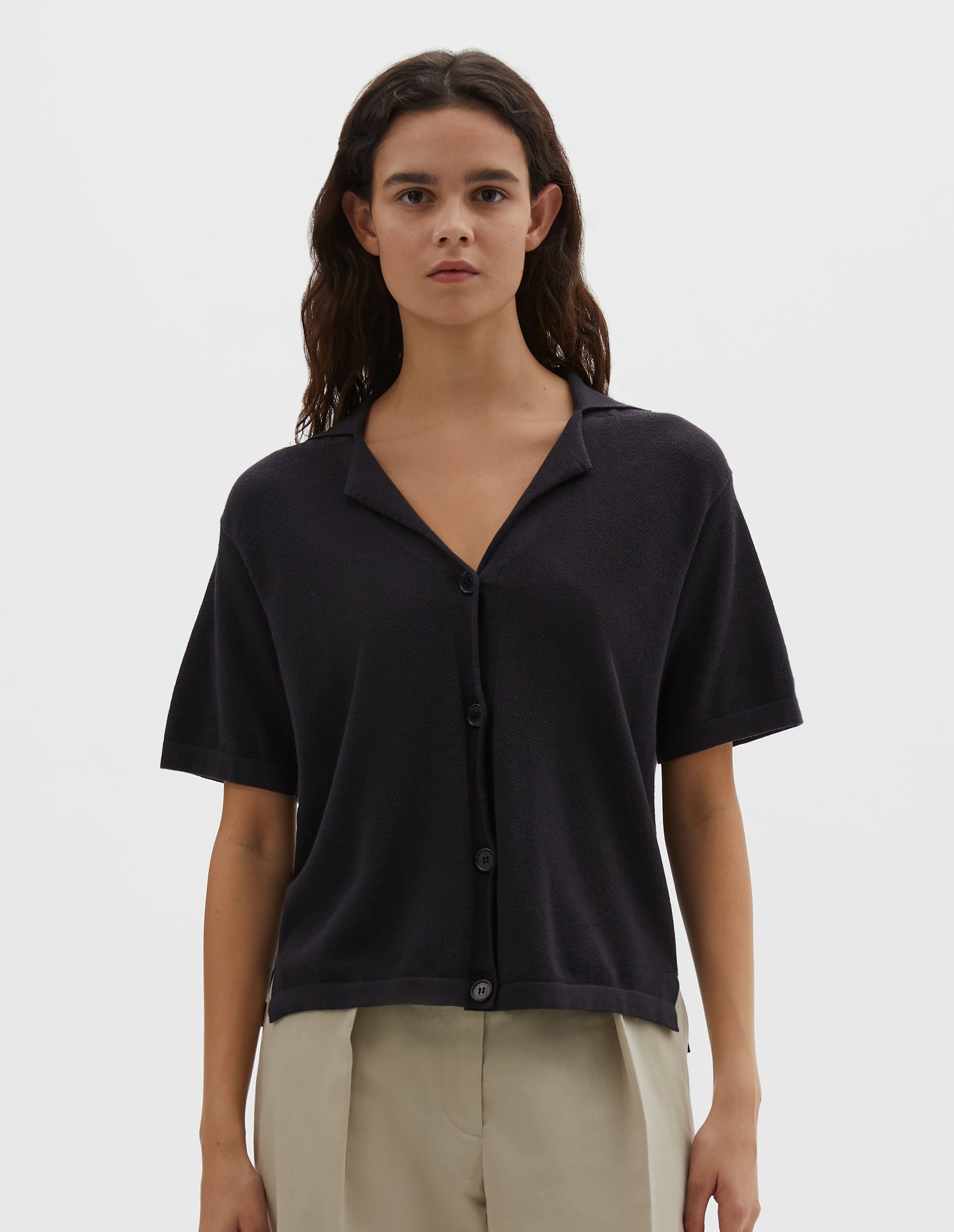 Cropped body image, front view of model wearing black knitted cotton short sleeve shirt, fine gauge rib collar, placket, cuffs and side split hem with horn buttons. Styled over cream pleated trousers.
