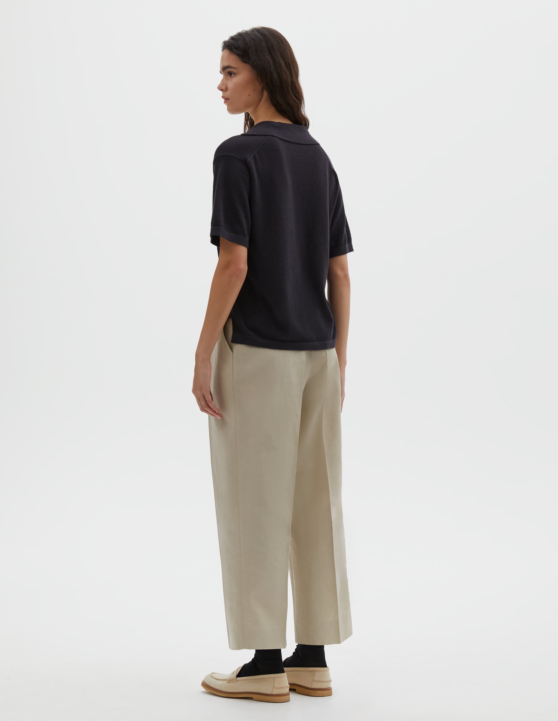 Full body image, back view of model wearing black knitted cotton short sleeve shirt, with fine gauge rib collar, placket, cuffs and side split hem. Styled over cream pleated trousers with black socks and cream shoes.