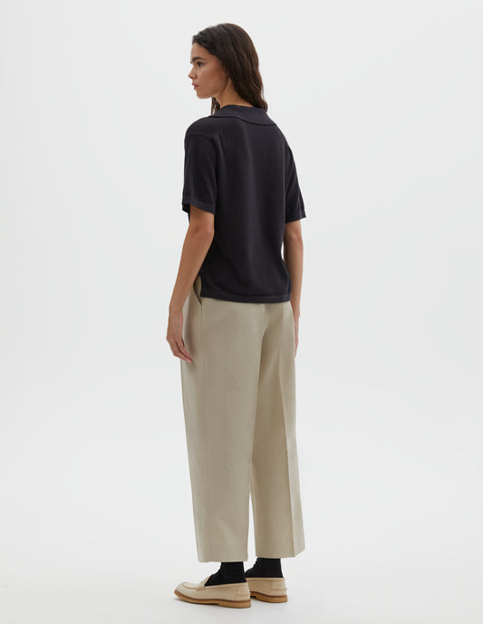 Full body image, back view of model wearing black knitted cotton short sleeve shirt, with fine gauge rib collar, placket, cuffs and side split hem. Styled over cream pleated trousers with black socks and cream shoes.