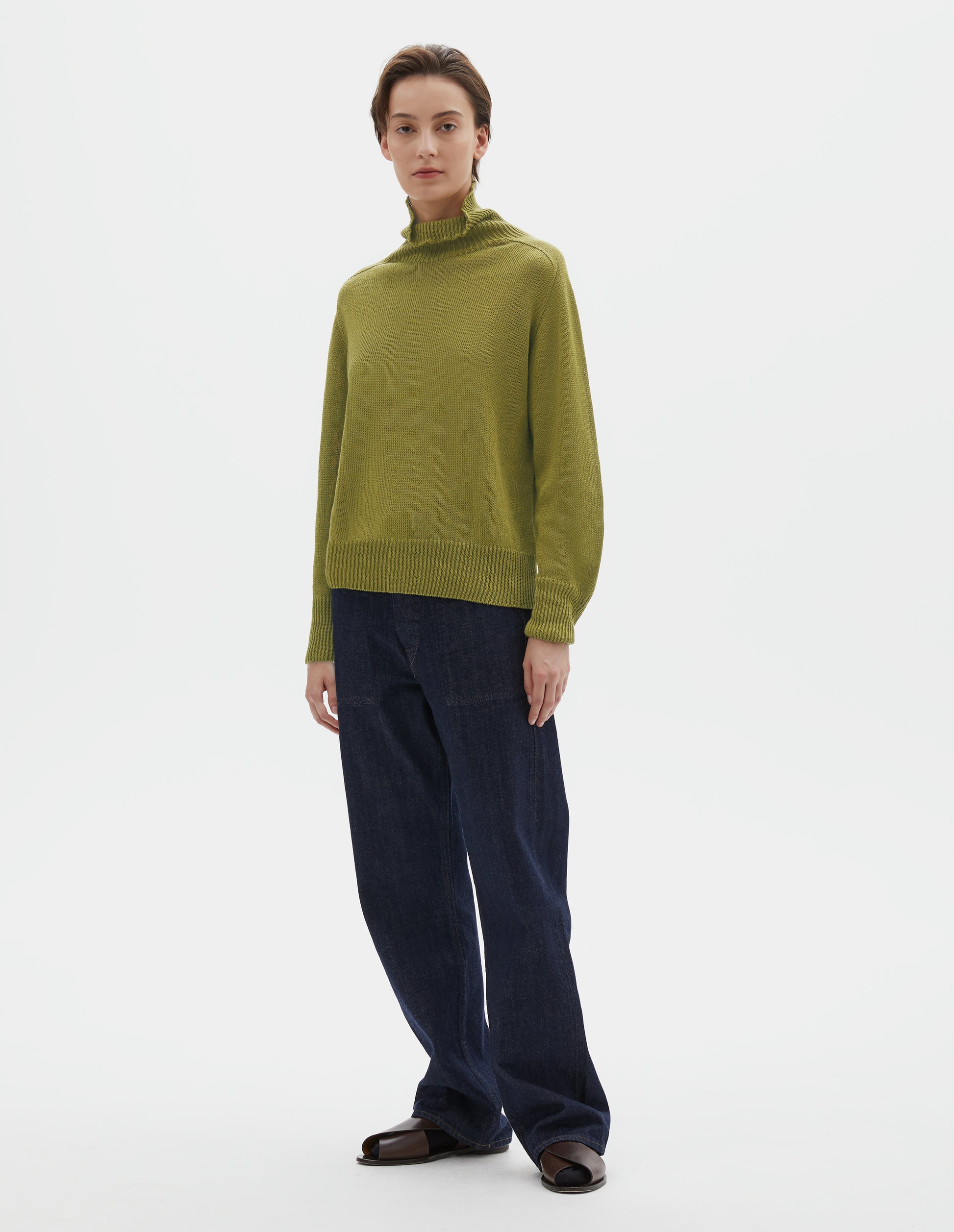 Full body image, front view of model wearing yellowish green linen cotton roll neck jumper with long saddle sleeves and contrast rib on neck, cuffs and hem. Styled with indigo blue jeans, black socks and brown leather sandals.