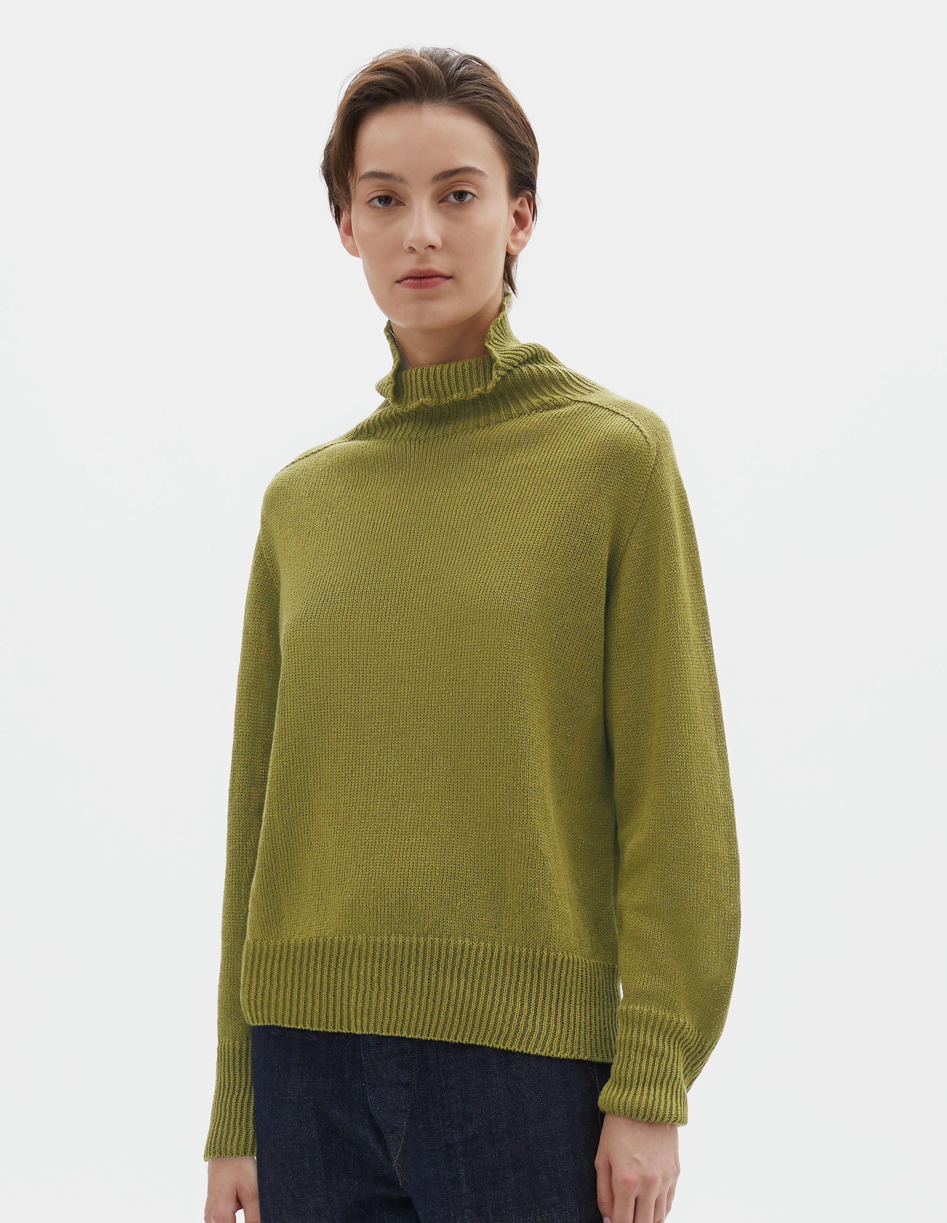 Cropped body image, front view of model wearing yellowish green linen cotton roll neck jumper with long saddle sleeves and contrast rib on neck, cuffs and hem. Styled with indigo blue jeans.