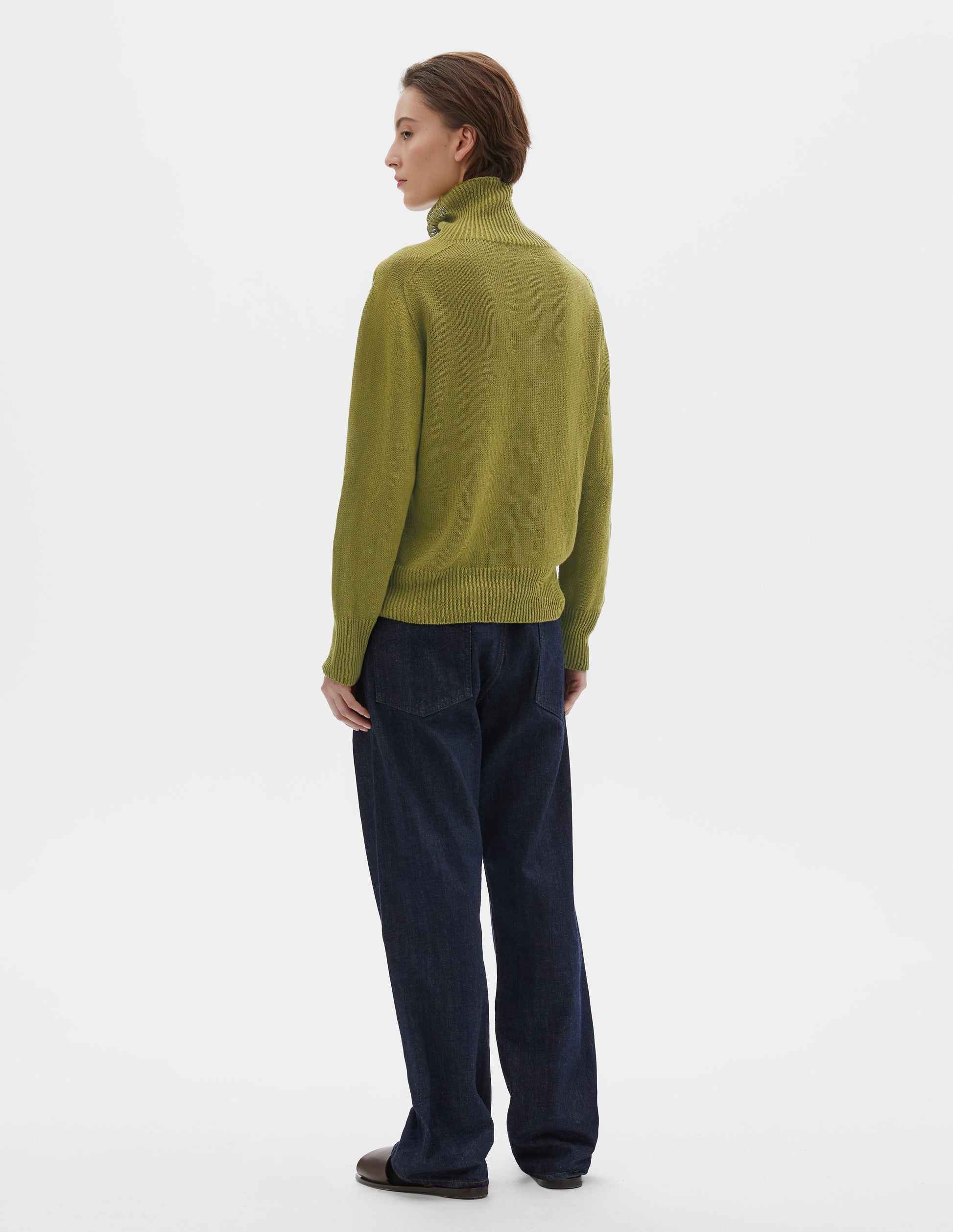 Full body image, back view of model wearing yellowish green linen cotton roll neck jumper with long saddle sleeves and contrast rib on neck, cuffs and hem. Styled with indigo blue jeans, black socks and brown leather sandals.