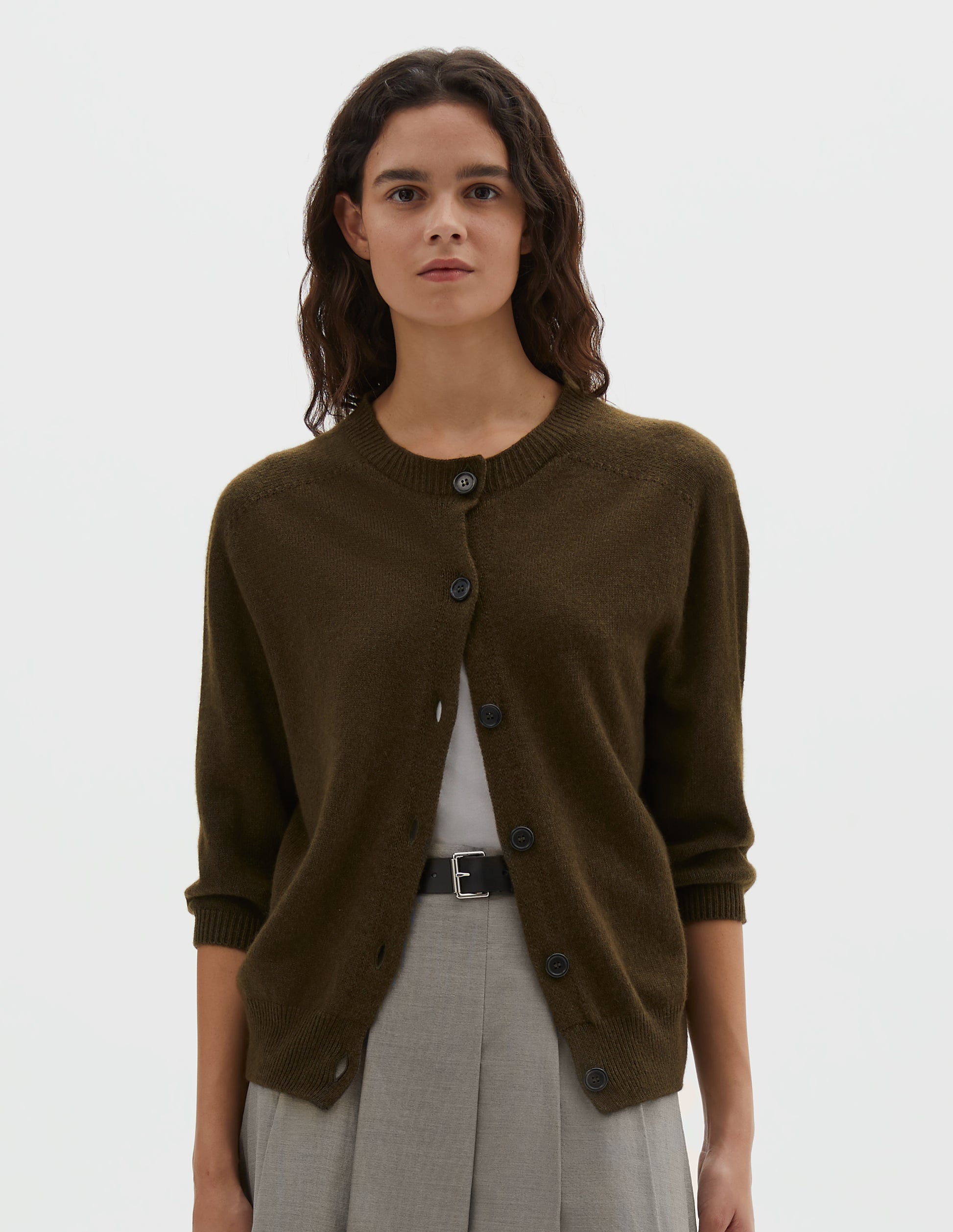 Cropped body image, front view of model wearing dark green cashmere cotton cardigan, with elbow length saddle sleeve and large horn buttons. Styled over white vest, with black leather belt and grey pleated skirt. 