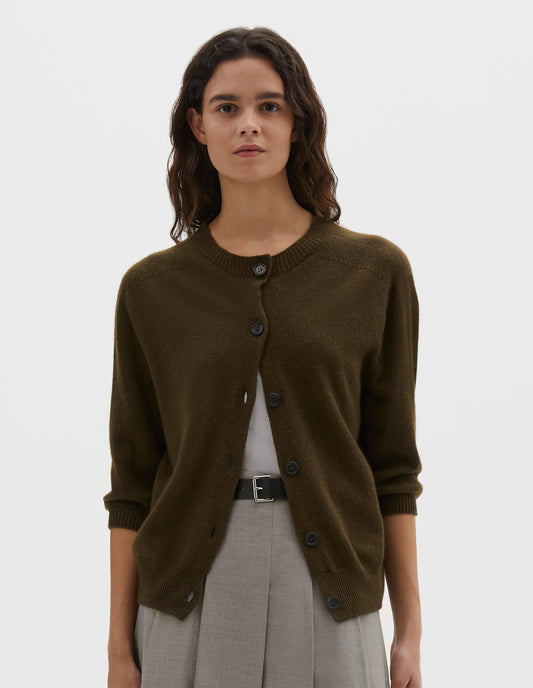 Cropped body image, front view of model wearing dark green cashmere cotton cardigan, with elbow length saddle sleeve and large horn buttons. Styled over white vest, with black leather belt and grey pleated skirt. 
