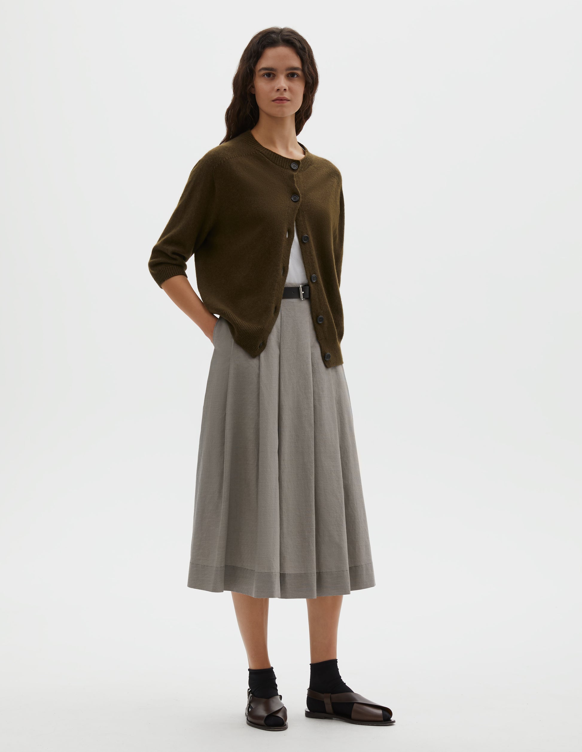 Full body image, front view of model wearing dark green cashmere cotton cardigan, with elbow length saddle sleeve and large horn buttons. Styled over white vest, with black leather belt, grey pleated skirt, black socks and brown leather sandals.