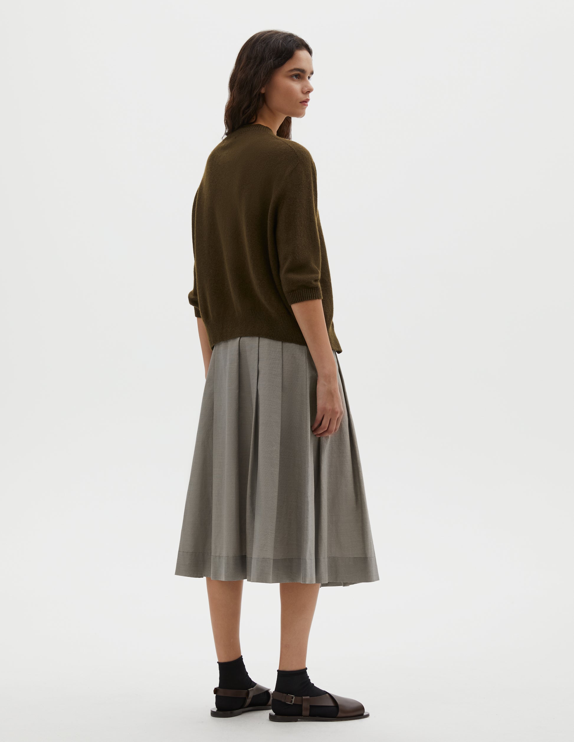 Full body image, back view of model wearing dark green cashmere cotton cardigan, with elbow length saddle sleeve. Styled with grey pleated skirt, black socks and brown leather sandals.