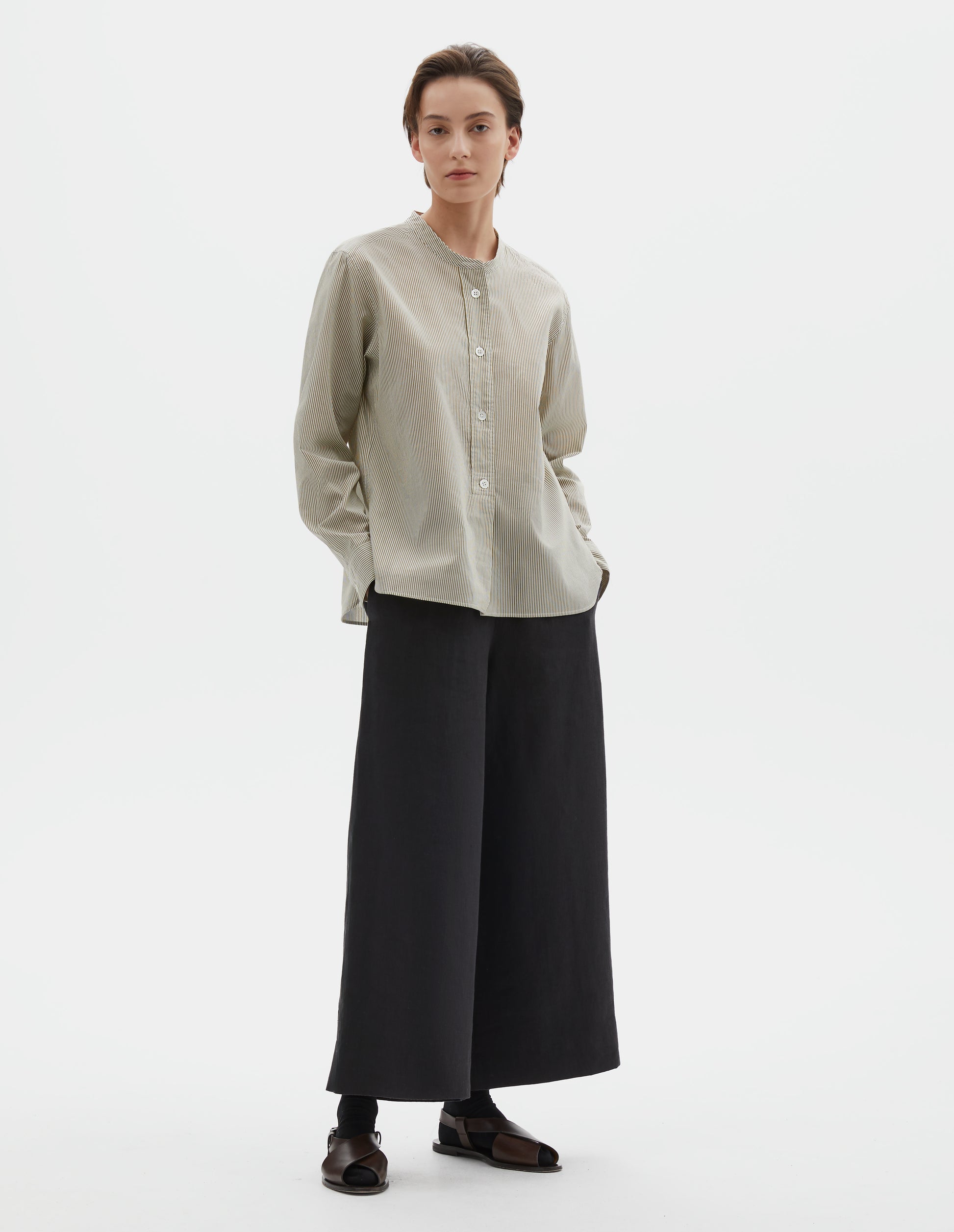 Full body image, front view of model wearing green and white striped cotton silk collarless shirt, with raised placket with box stitch detailing and horn button closure. Styled with black culotte trousers, black socks and brown leather sandals.