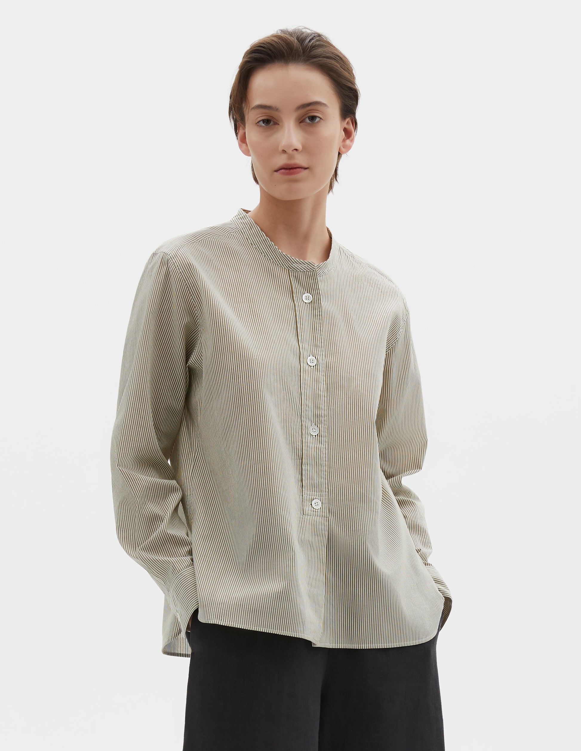 Cropped body image, front view of model wearing green and white striped cotton silk collarless shirt, with raised placket with box stitch detailing and horn button closure. Styled with black culotte trousers.