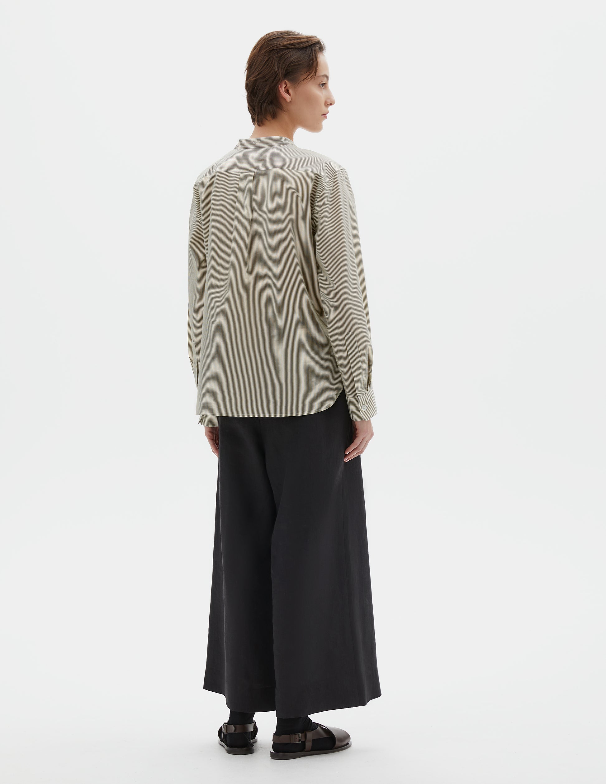 Full body image, back view of model wearing green and white striped cotton silk collarless shirt, with box pleat on back yoke, elongated back hem and horn button cuffs. Styled with black culotte trousers, black socks and brown leather sandals. 