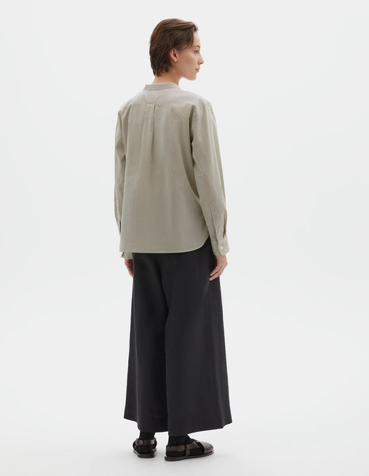 Full body image, back view of model wearing green and white striped cotton silk collarless shirt, with box pleat on back yoke, elongated back hem and horn button cuffs. Styled with black culotte trousers, black socks and brown leather sandals. 