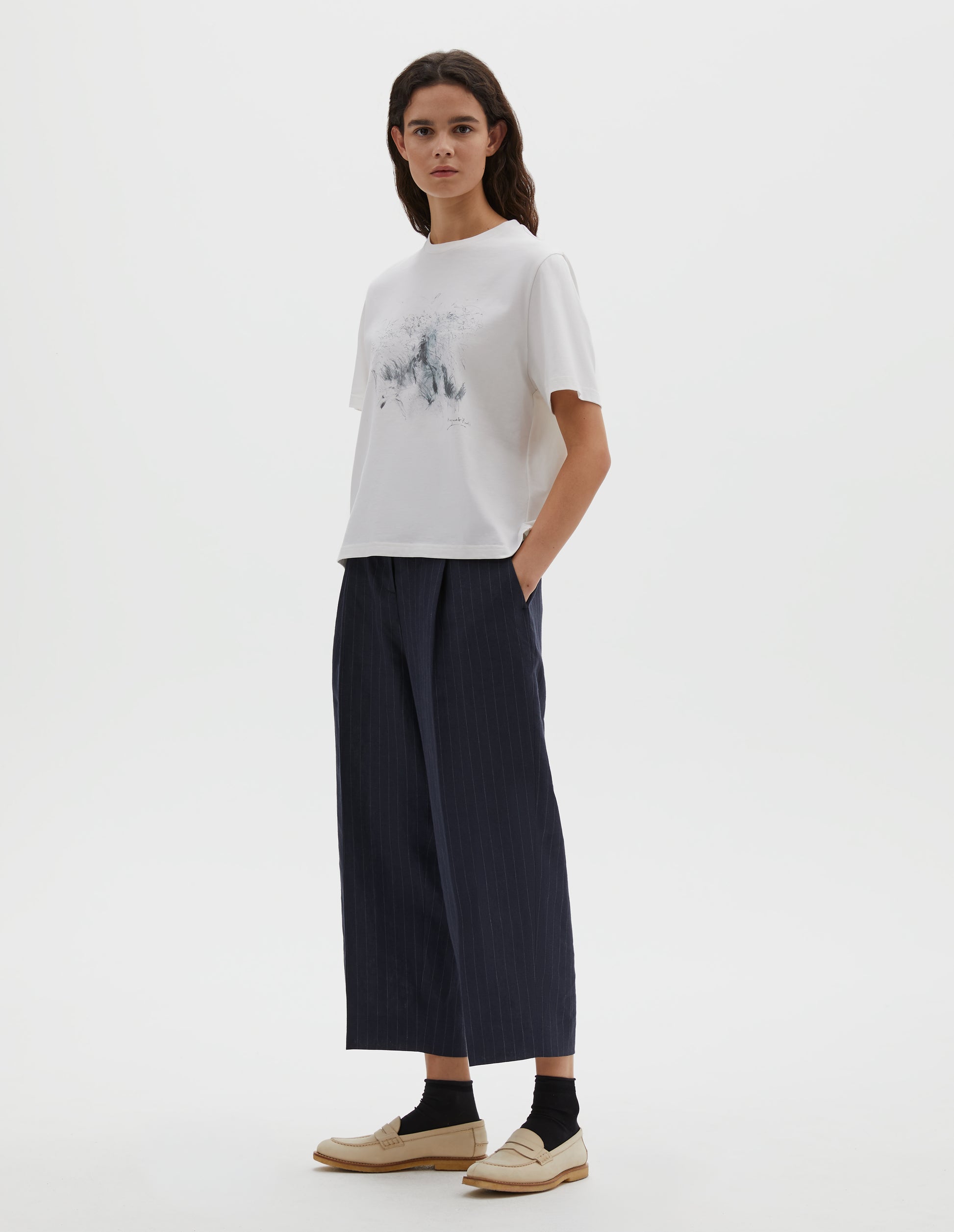 Full body image, front view of model wearing white relaxed, boxy crew neck t-shirt with dark grey drawing print. Styled with dark blue striped trousers, black socks and beige leather loafers. 