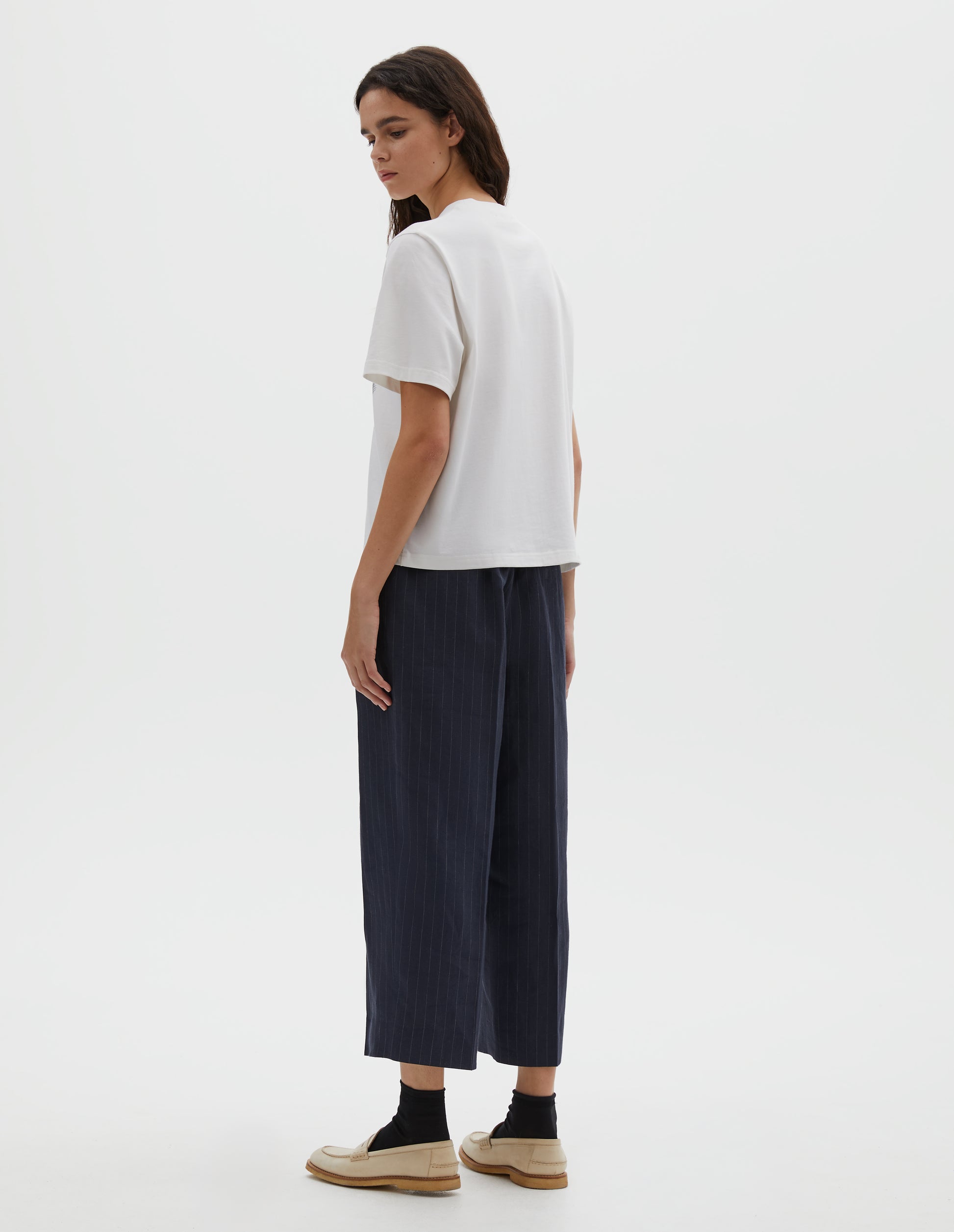 Full body image, back view of model wearing white relaxed, boxy crew neck t-shirt. Styled with dark blue striped trousers, black socks and beige leather loafers. 