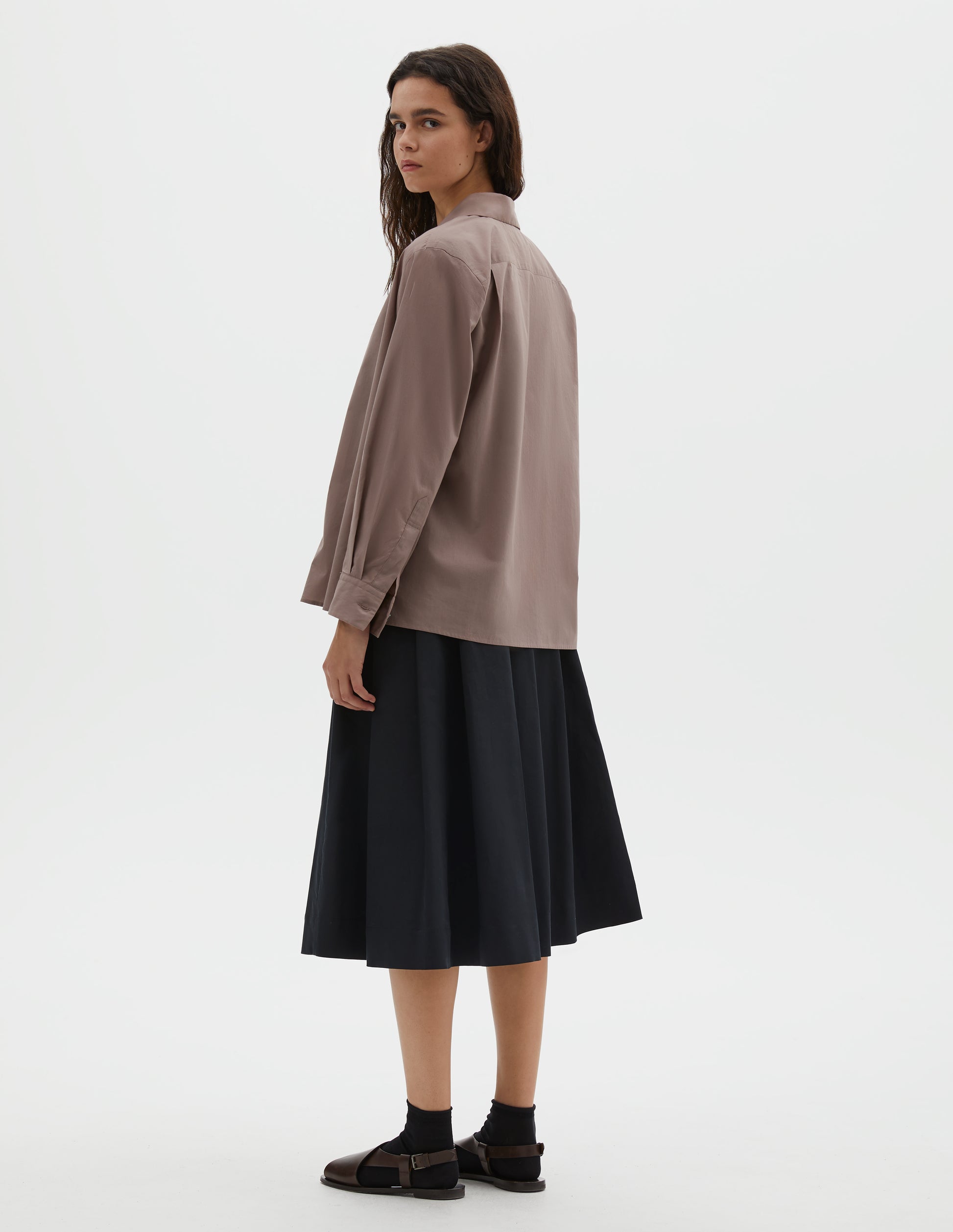 Full body image, back view of model wearing pinkish brown shirt with pleats on the back yoke, and a side split hem. Styled with blue skirt and black socks with dark brown leather sandals.