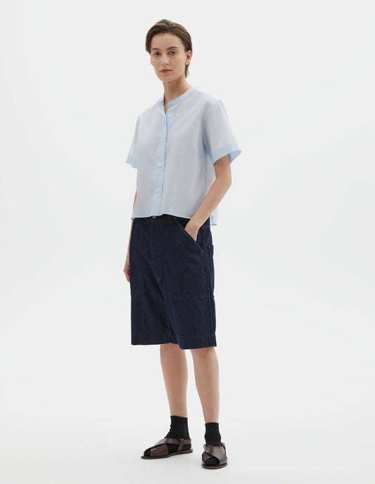 Full body image, front view of model wearing regular cut cropped light blue collarless shirt with short sleeves, side split stepped hem and mother of pearl buttons on raised placket. Styled with dark blue denim shorts, black socks and dark brown leather sandals.