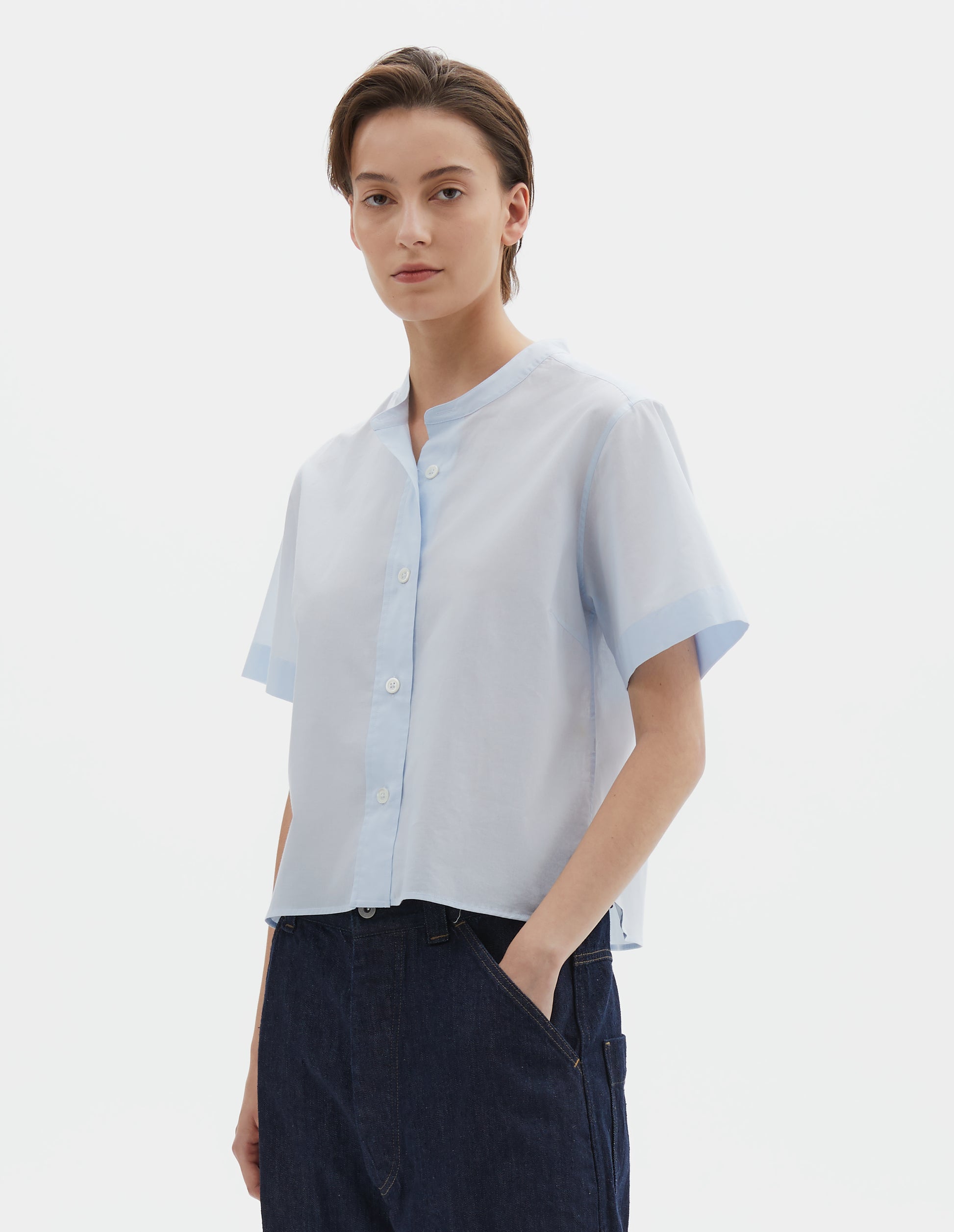 Cropped body image, front view of model wearing regular cut cropped light blue collarless shirt with short sleeves, side split stepped hem and mother of pearl buttons on raised placket. Styled with dark blue denim shorts.