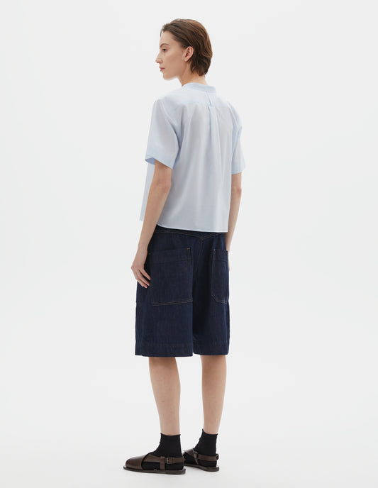 Full body image, back view of model wearing regular cut cropped light blue collarless shirt with short sleeves, side split stepped hem, box pleat and hanger loop detail on back yoke, and buttonhole detailing on collar band. Styled with dark blue denim shorts, black socks and dark brown leather sandals.