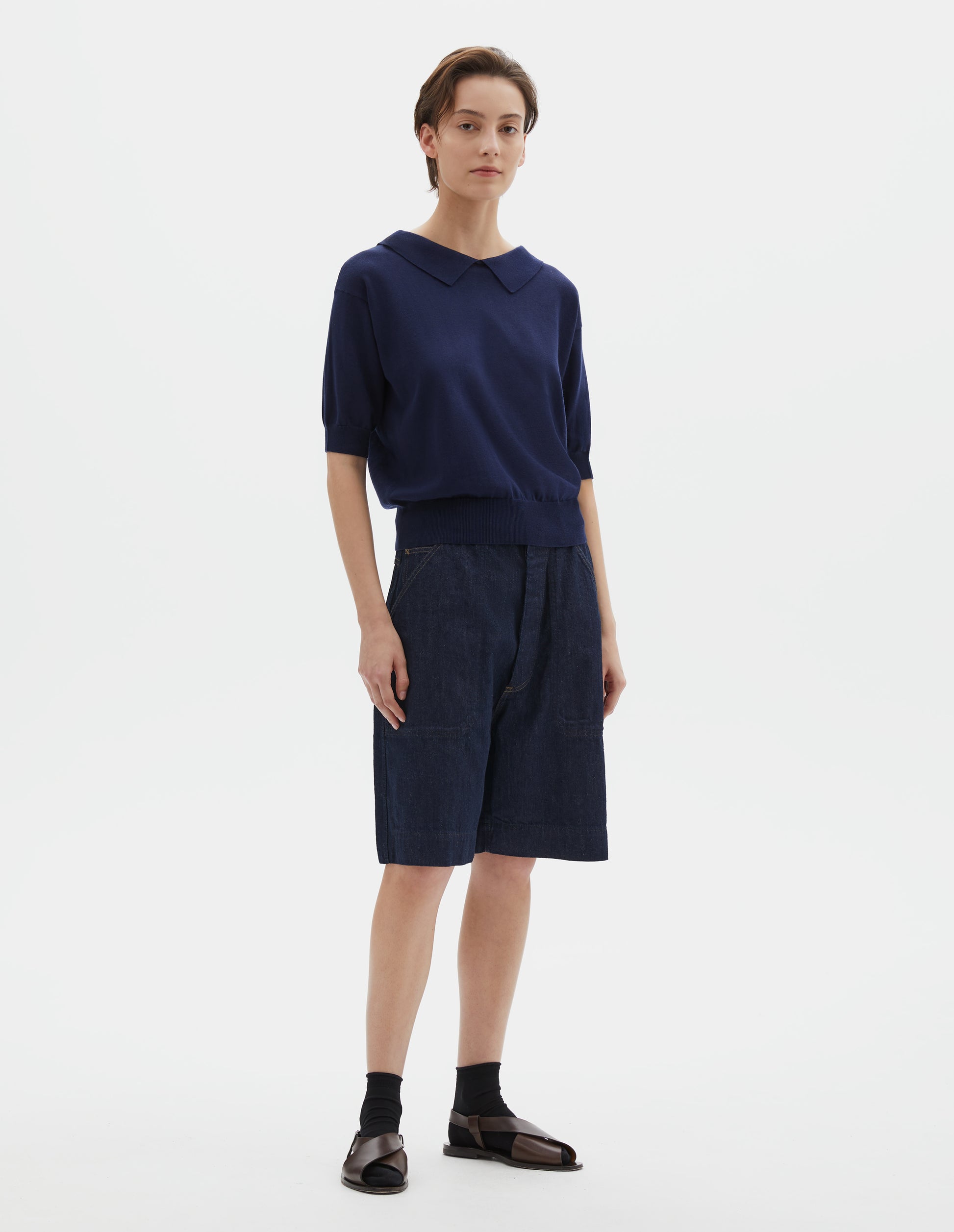 Full body image, front view of model wearing dark navy cotton and linen knitted t-shirt, with cutaway collar and rib cuff and hem. Styled with indigo blue denim shorts, black ankle socks and brown leather sandals.