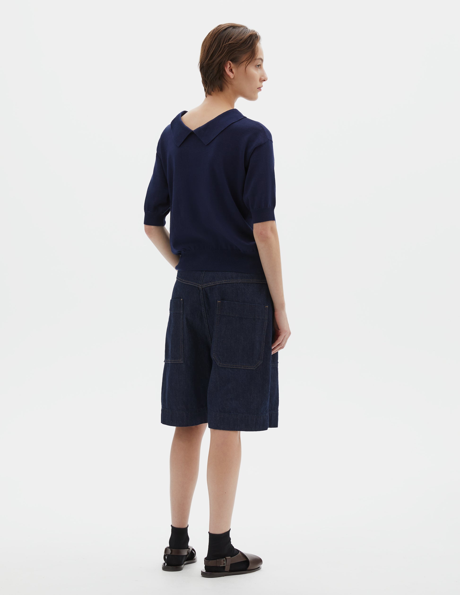 Full body image, back view of model wearing dark navy cotton and linen knitted t-shirt, with cutaway collar and rib cuff and hem. Styled with indigo blue denim shorts, black ankle socks and brown leather sandals.