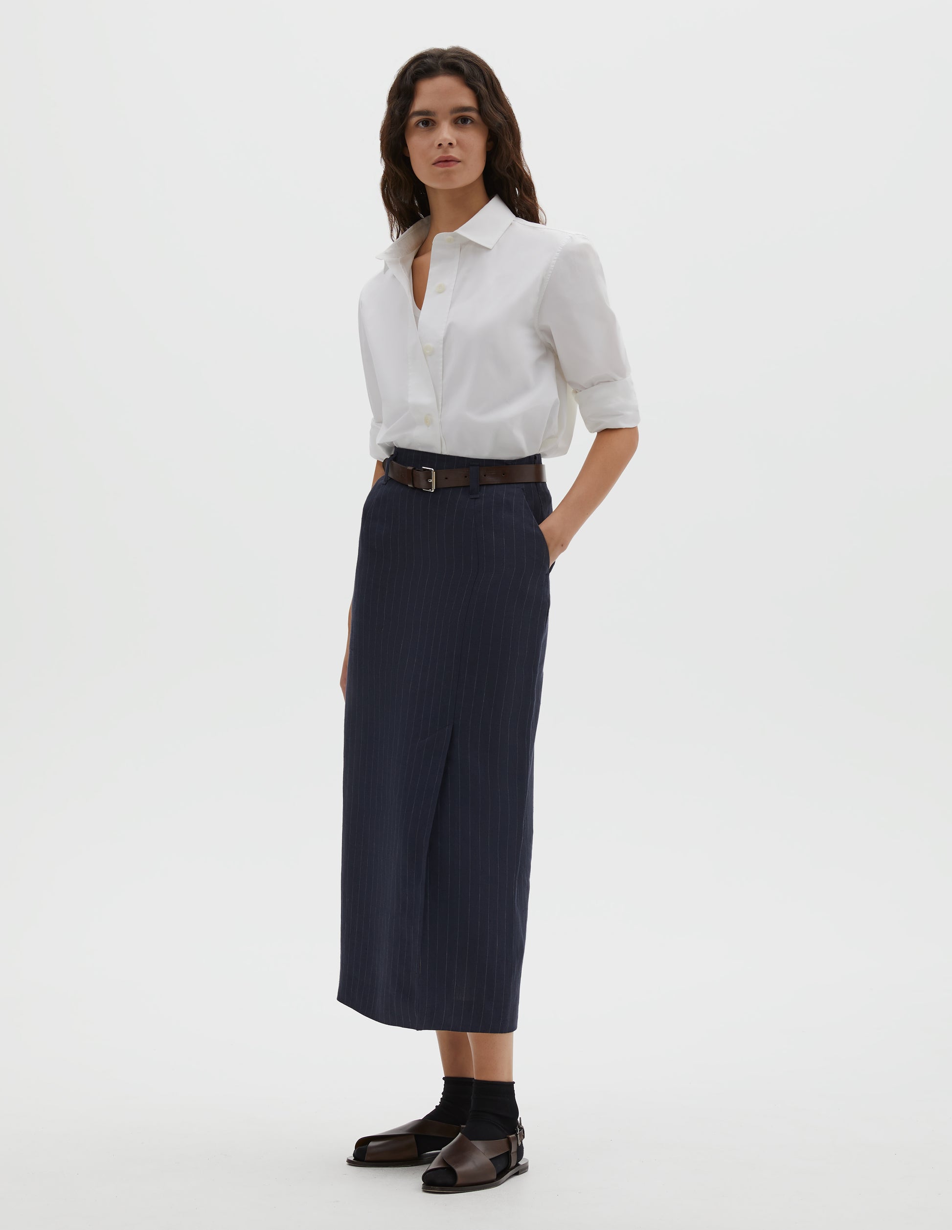 Full body image, front view of model wearing navy and grey pinstripe linen skirt, with offset front vent, concealed side zip fastening, slant side pockets and belt loops. Styled with white cotton shirt tucked in, with brown leather belt, black ankle socks and brown leather sandals.