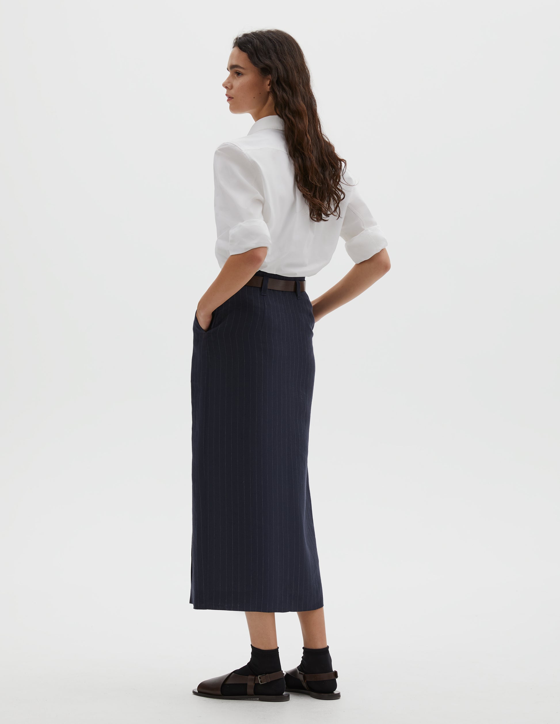 Full body image, back view of model wearing navy and grey pinstripe linen skirt, with belt loops. Styled with white cotton shirt tucked in, with brown leather belt, black ankle socks and brown leather sandals.