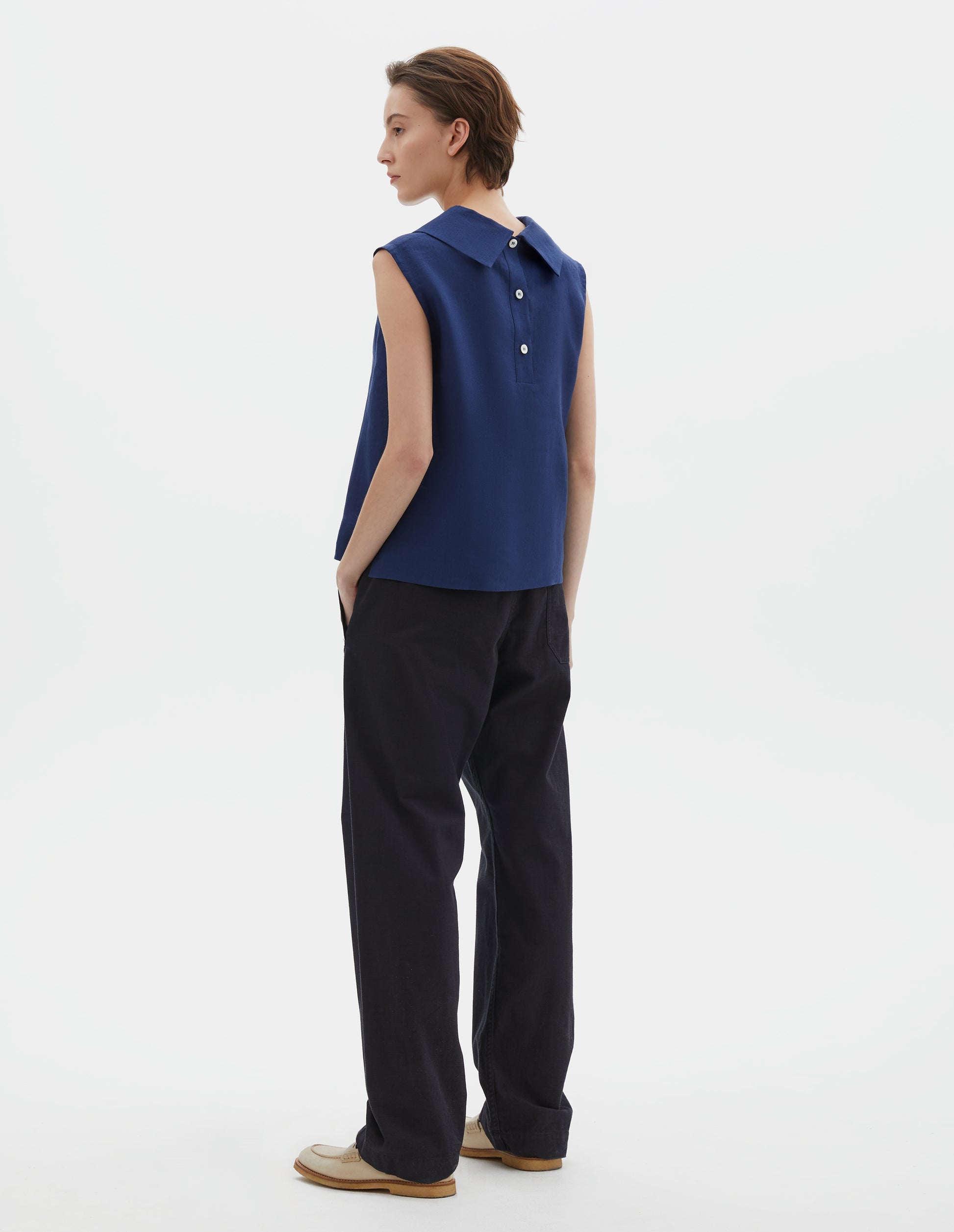 Full body image, back view of model wearing dark blue regular cut Irish linen top with exaggerated collar, button-fastening placket at the centre back, with a deep hem with side splits. Styled with dark blue trousers.
