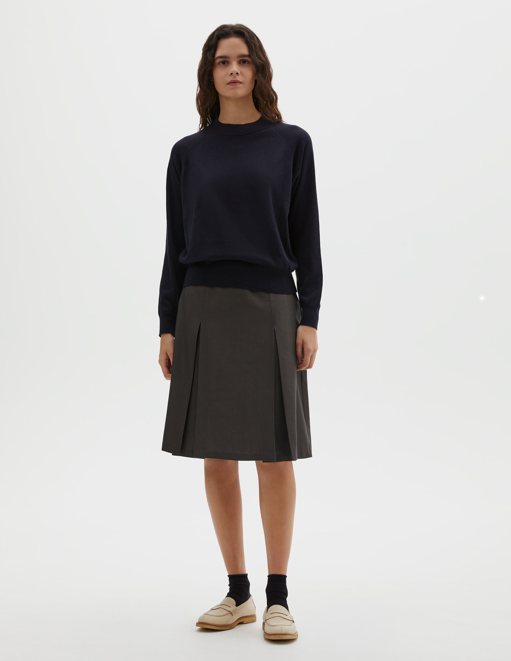 Full body image, front view of model wearing dark navy merino wool and cotton long sleeve jumper with raglan sleeve, deep rib crew neck and single rib cuffs and hem. Styled with pleated dark green skirt, black ankle socks and beige suede loafers.