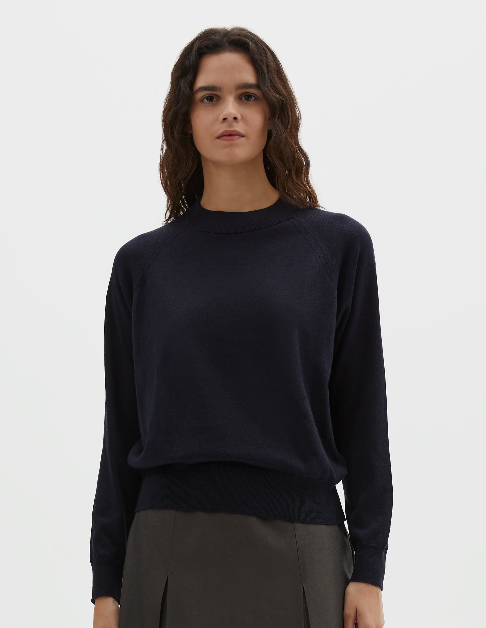 Cropped body image, front view of model wearing dark navy merino wool and cotton long sleeve jumper with raglan sleeve, deep rib crew neck and single rib cuffs and hem. Styled with pleated dark green skirt.