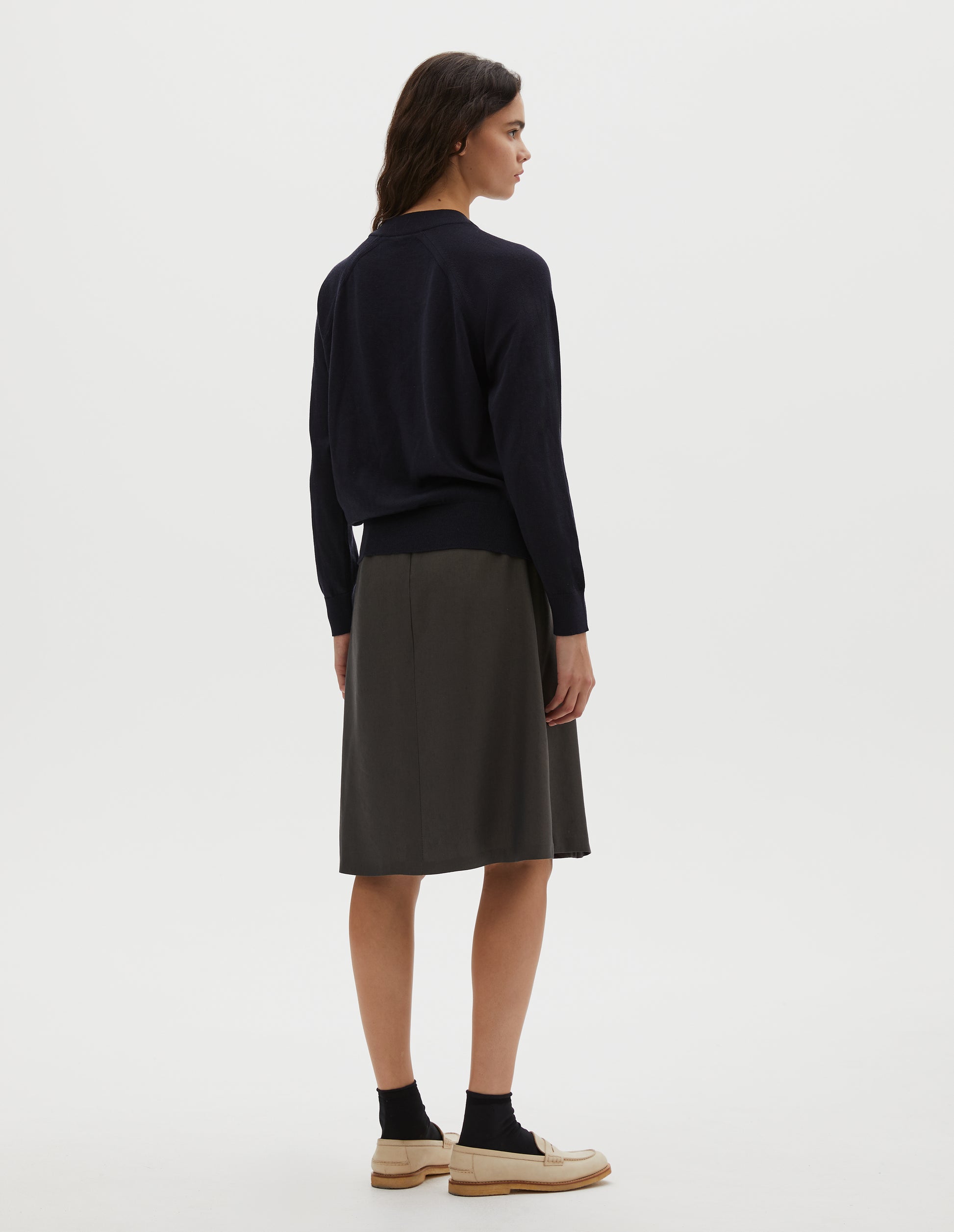 Full body image, back view of model wearing dark navy merino wool and cotton long sleeve jumper with raglan sleeve, deep rib crew neck and single rib cuffs and hem. Styled with pleated dark green skirt, black ankle socks and beige suede loafers.