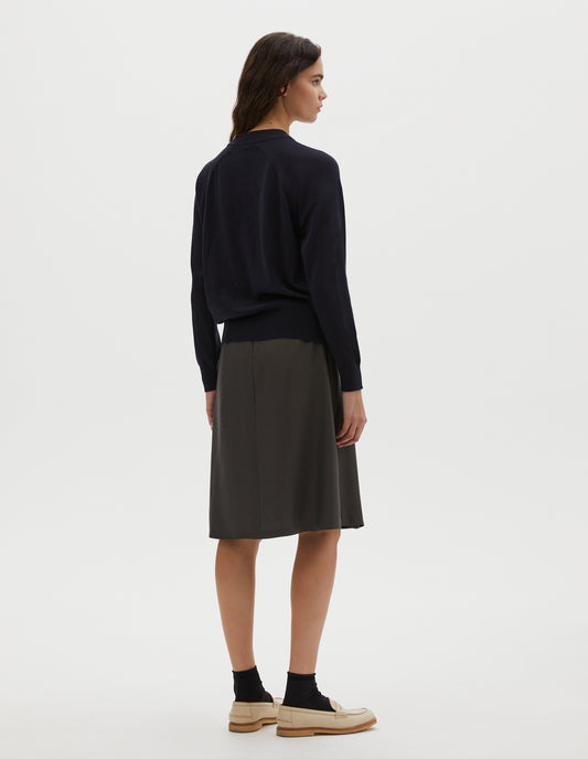 Full body image, back view of model wearing dark navy merino wool and cotton long sleeve jumper with raglan sleeve, deep rib crew neck and single rib cuffs and hem. Styled with pleated dark green skirt, black ankle socks and beige suede loafers.