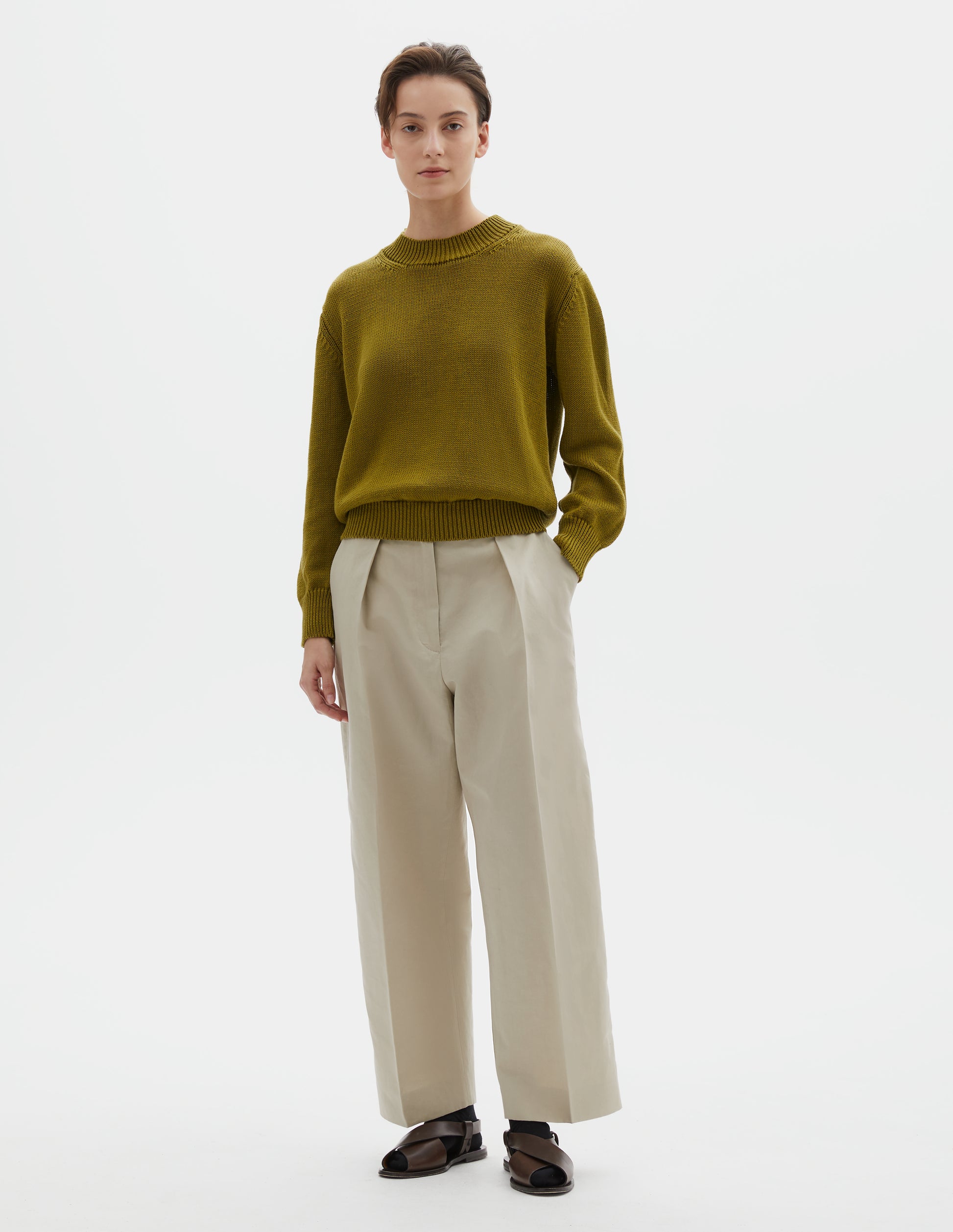 Full body image, front view of model wearing greenish yellow linen and cotton crew neck jumper, with deep rib crew neck, rib cuffs and hem. Styled with beige linen trousers, black socks and brown leather sandals.