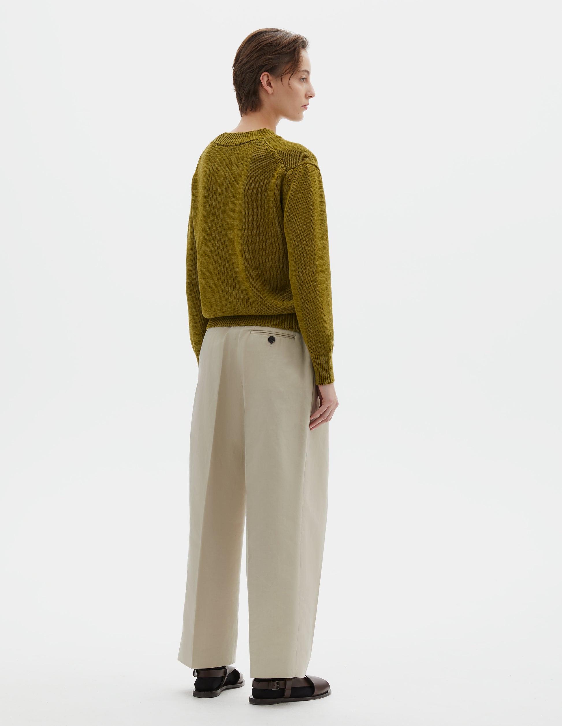 Full body image, back view of model wearing greenish yellow linen and cotton crew neck jumper, with deep rib crew neck, rib cuffs and hem. Styled with beige linen trousers, black socks and brown leather sandals.