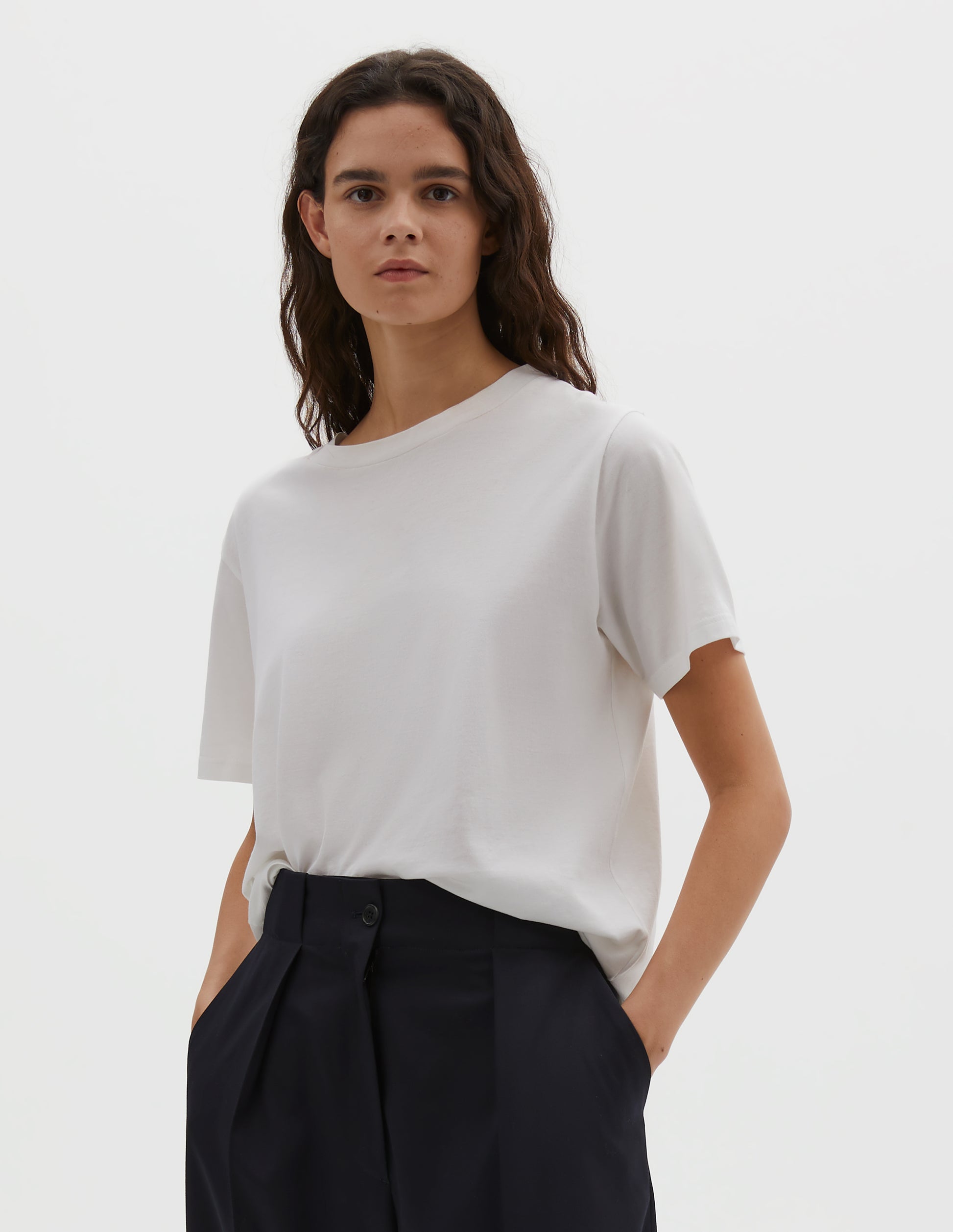 Cropped body image, front view of model wearing off white cotton short sleeve t-shirt with boxy cut, Styled with dark navy pleated trousers.